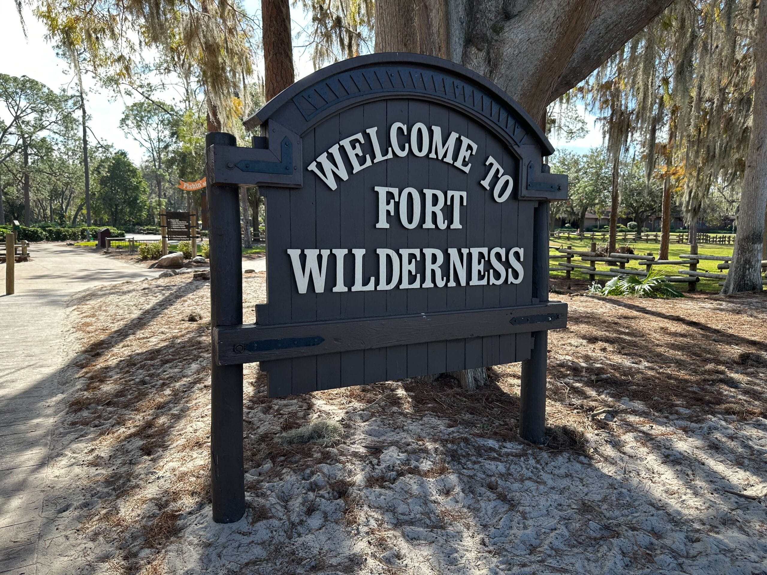 A wooden sign reads "Welcome to Fort Wilderness" set against a backdrop of trees and a sandy path.