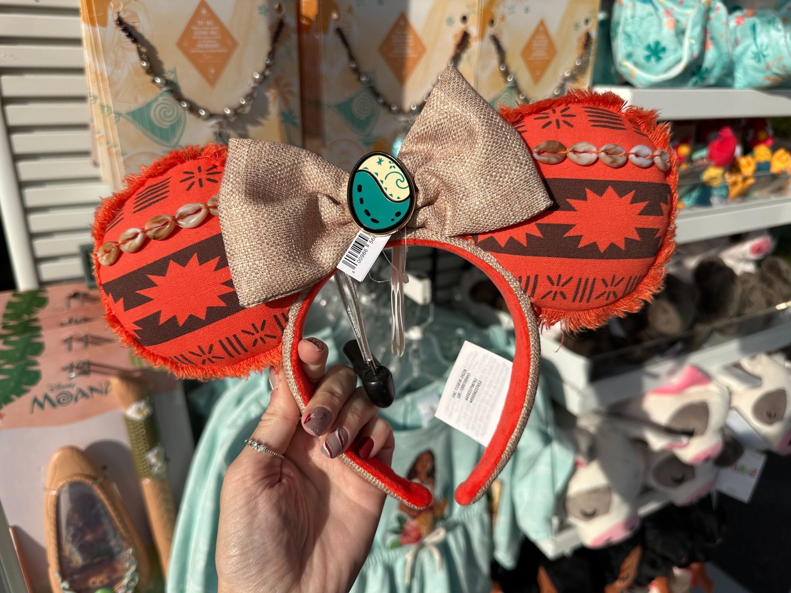 A hand holds a pair of mouse ears headband with a tribal pattern and a bow, in a store display with various plush toys and accessories in the background.