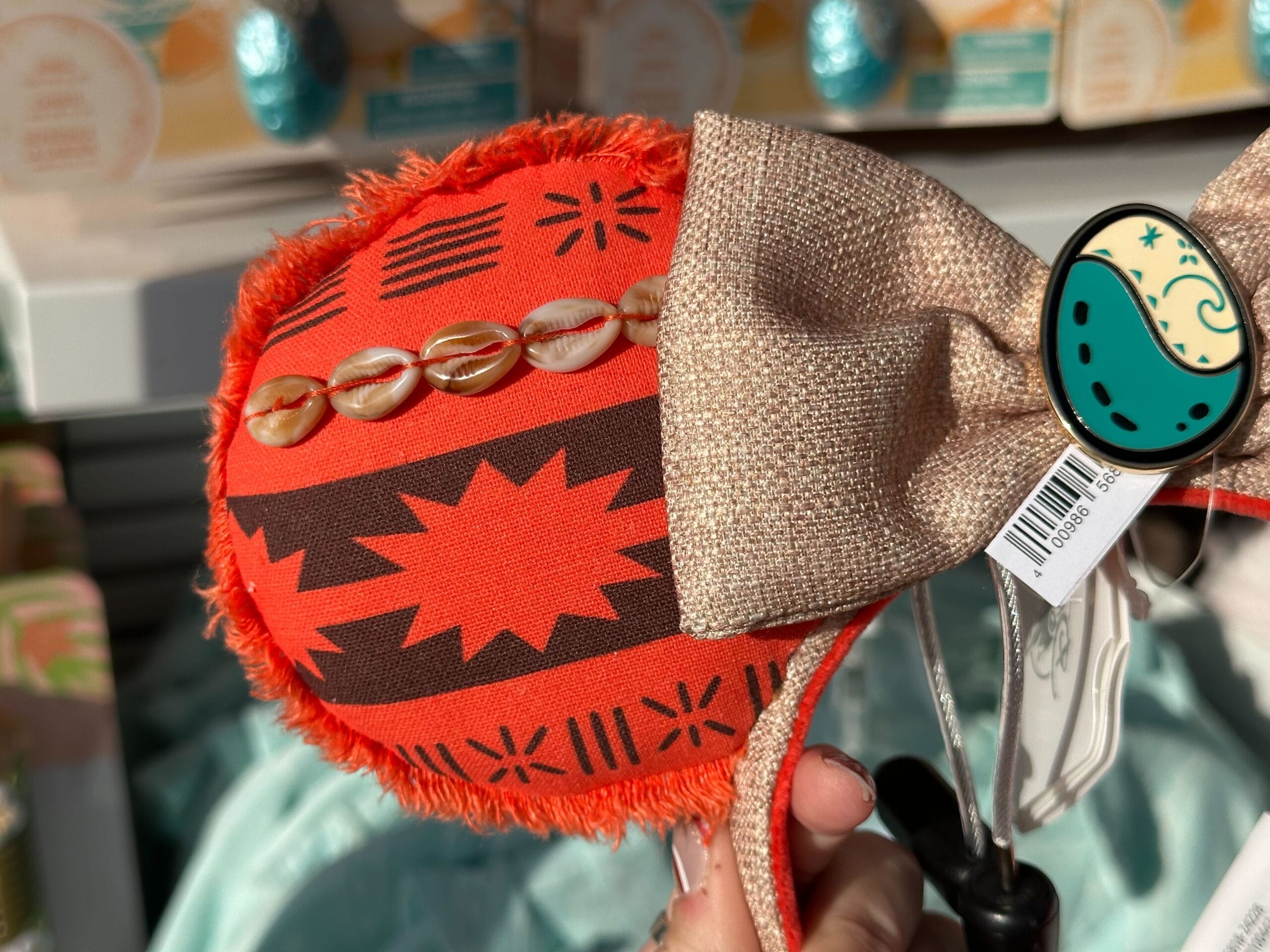 Close-up of a headband with orange fabric featuring tribal patterns and cowrie shells, adorned with a large gold bow and a decorative pin.