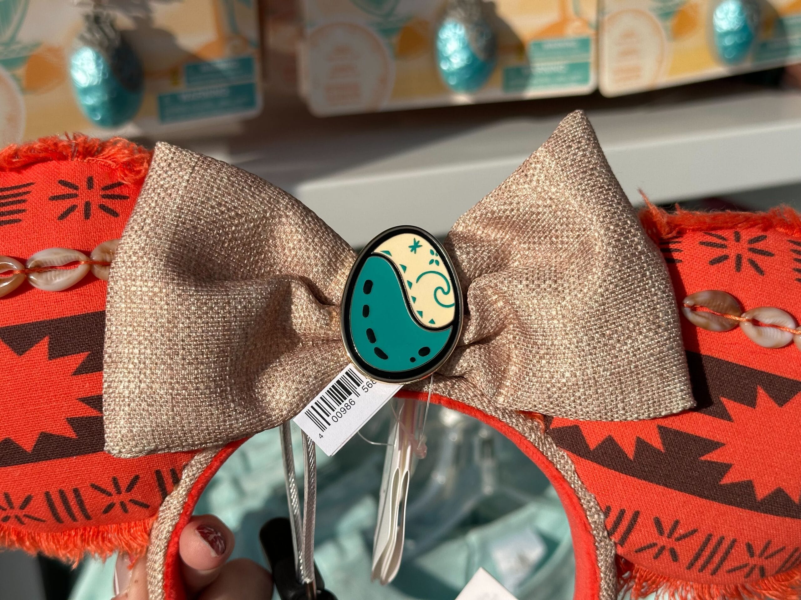 A pair of ears headband with an orange and black tribal pattern, topped with a beige bow. The center features an oval emblem with a green and cream design. Tags are attached.