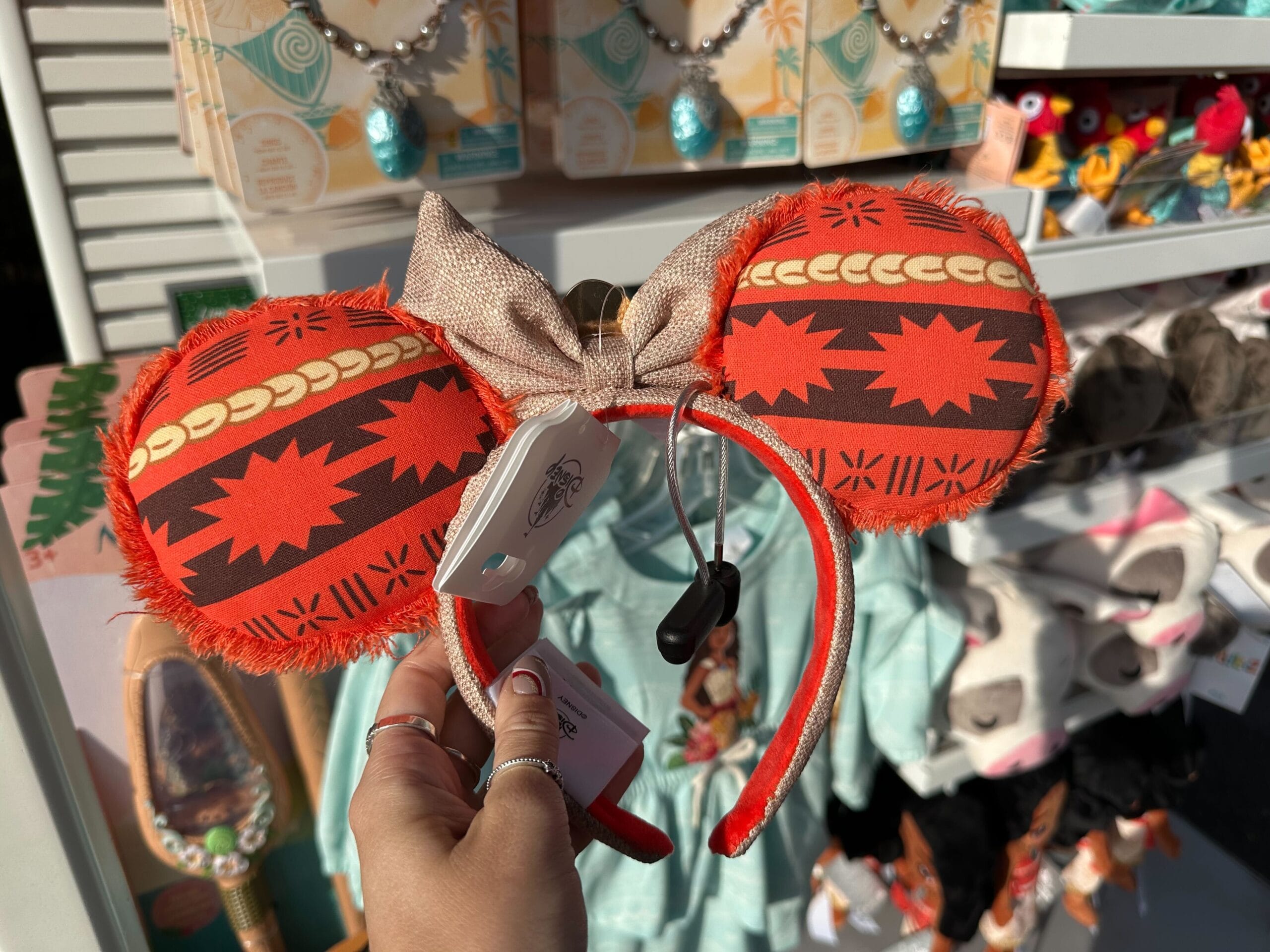 A person holds red and brown patterned Minnie Mouse ears with a gold bow in a store display of various themed merchandise.
