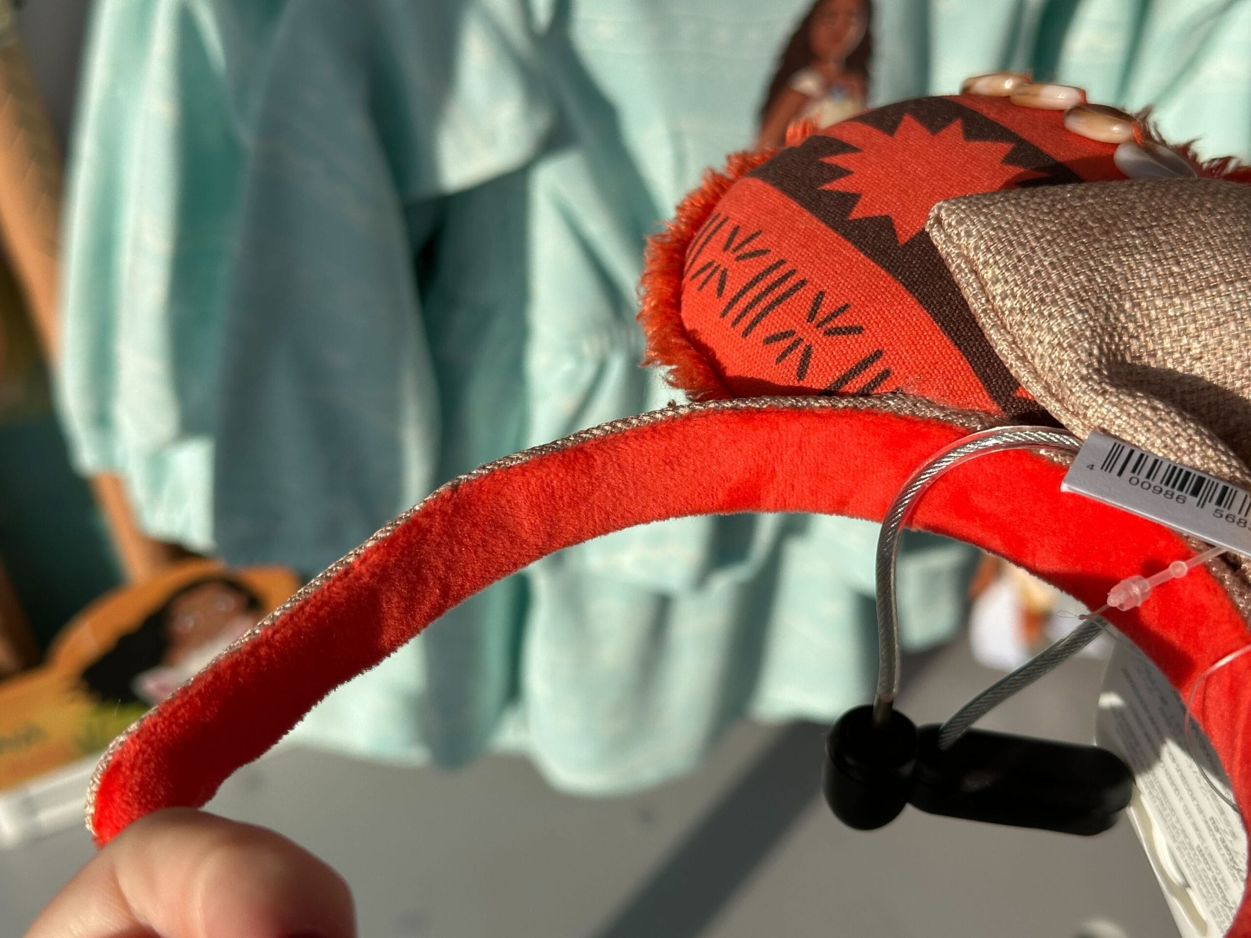 Close-up of a hand holding a red headband decorated with patterned fabric and a bow. A teal garment is visible in the background.