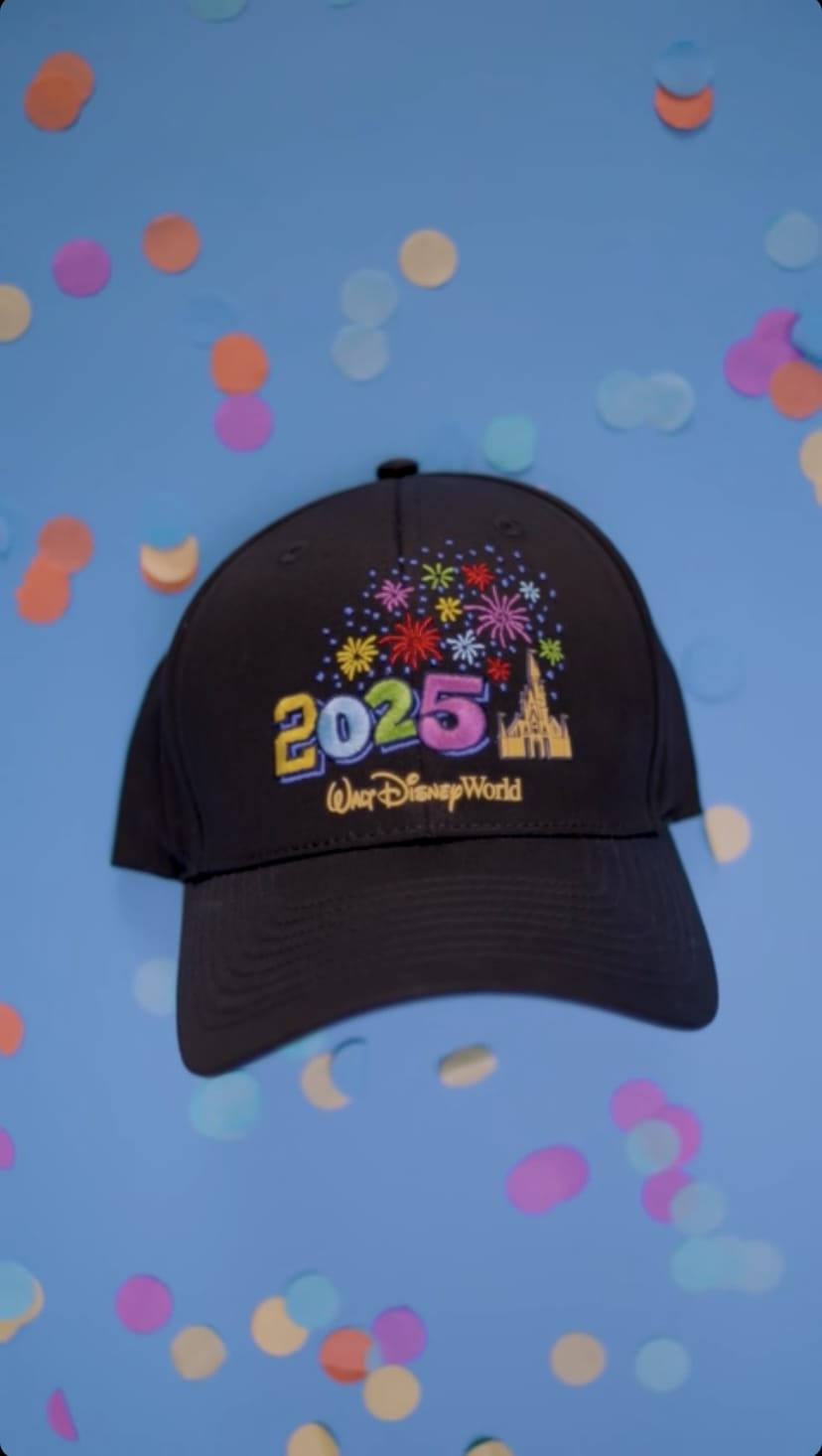 A black Disney World cap featuring "2025" with fireworks design and a castle, set against a blue background with colorful confetti.