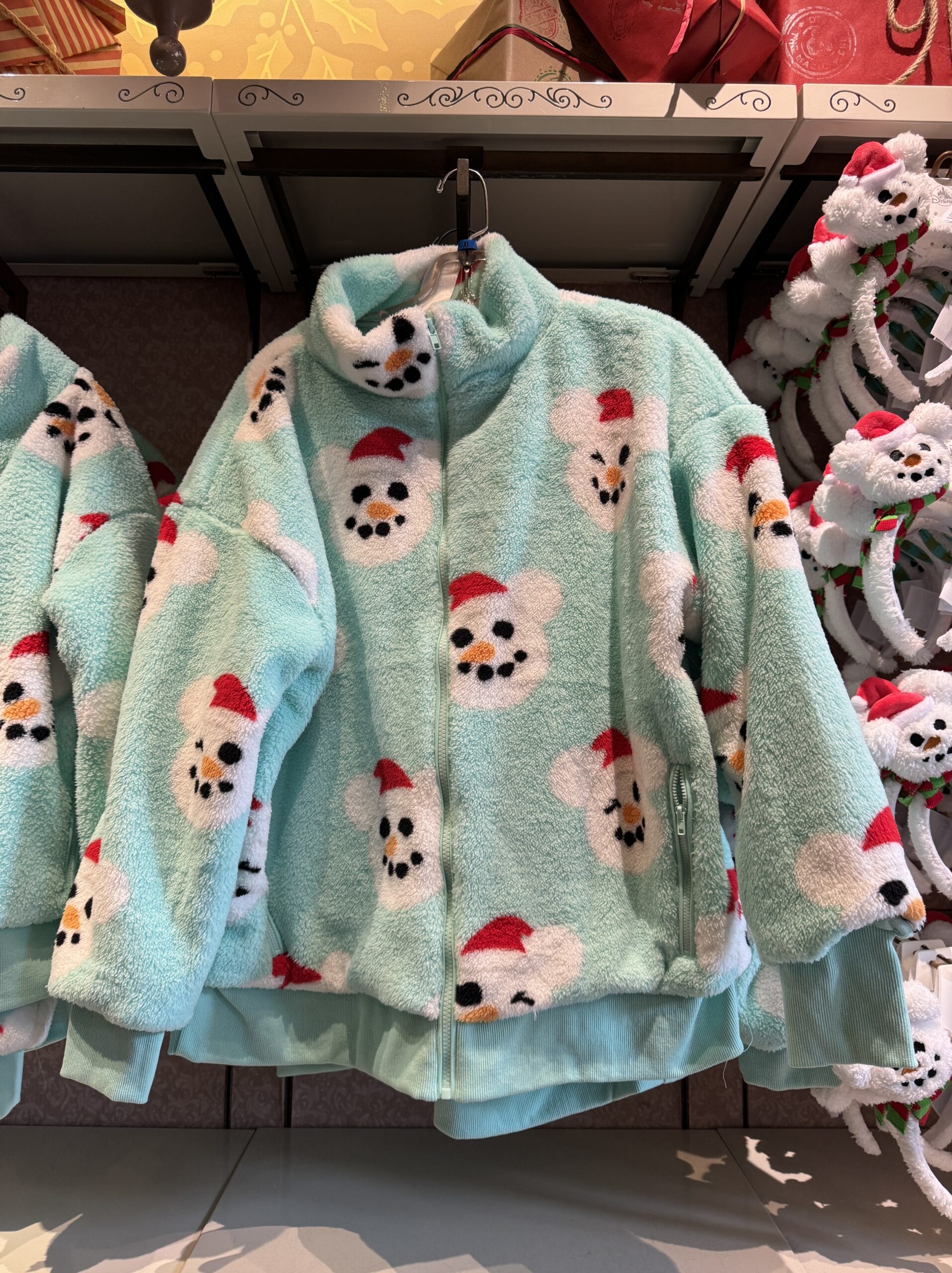 A light blue fleece jacket with a pattern of snowman faces wearing red hats is charmingly displayed in the store, evoking the whimsical spirit of Magic Kingdom.