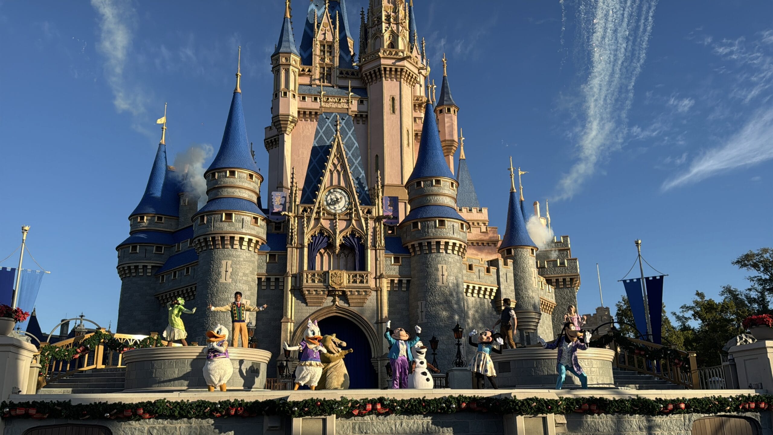 Characters perform in front of the Magic Kingdom's castle with blue spires and pink accents, under a clear sky with contrails.