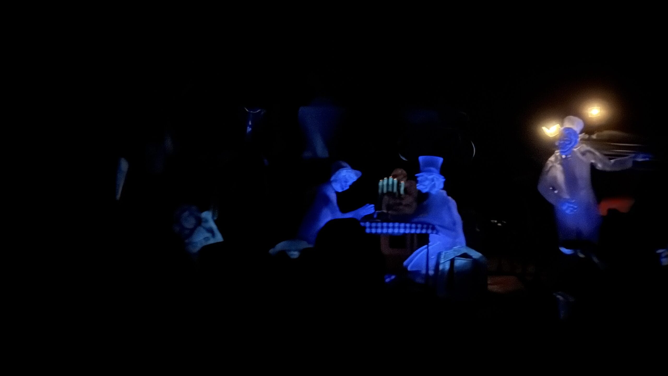 In a dimly lit scene, two ghostly figures in blue light sit at a table with a cake, creating an otherworldly atmosphere straight out of the Magic Kingdom. This enchanting moment, captured in our 2024 PHOTO REPORT, enchants viewers with its mystical charm.