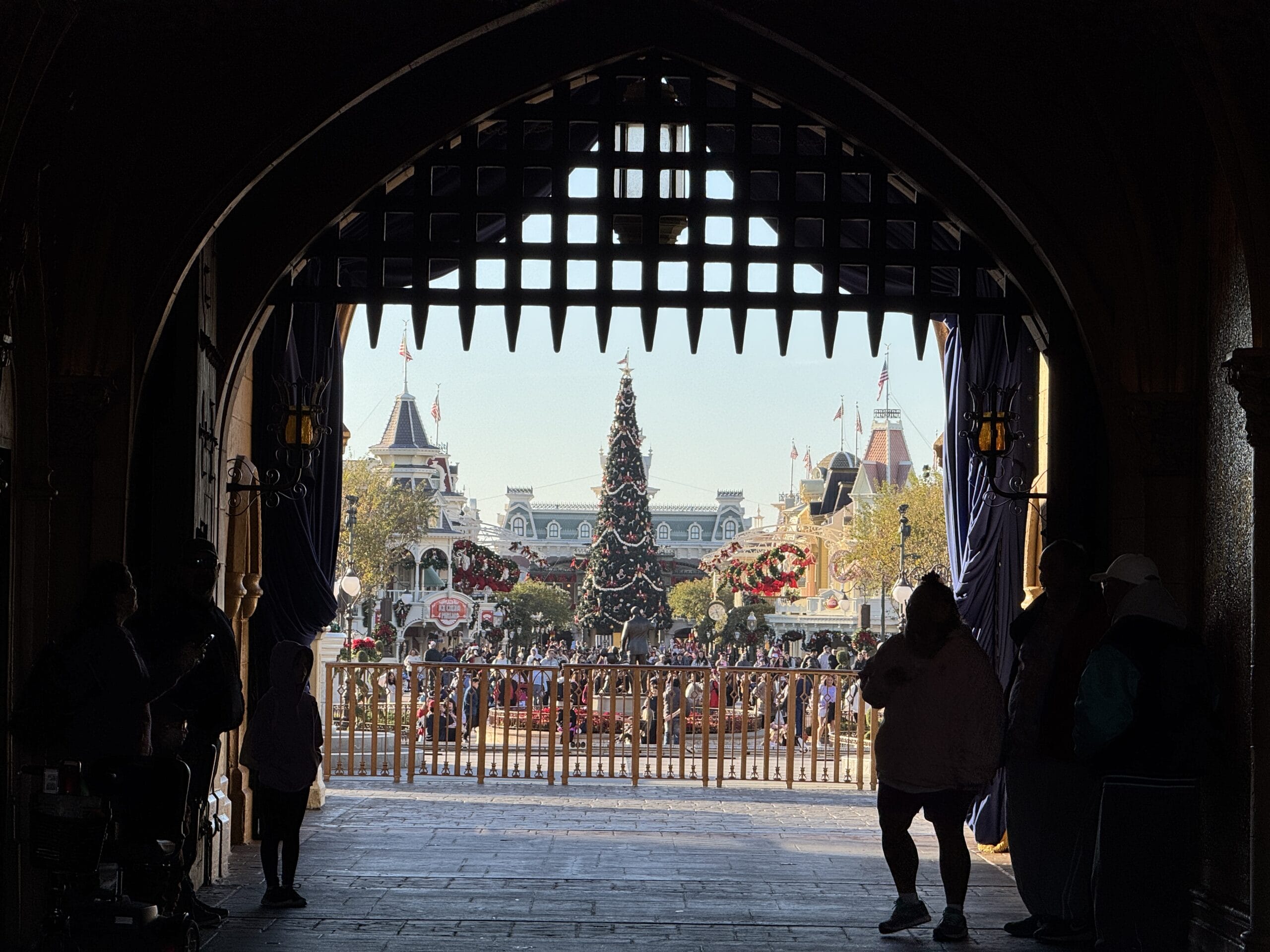 View from inside a castle archway looking out towards a festive plaza in the Magic Kingdom, with a large Christmas tree and people walking around. Perfect scene for a 2024 holiday photo report, capturing the enchanting joy of the season.