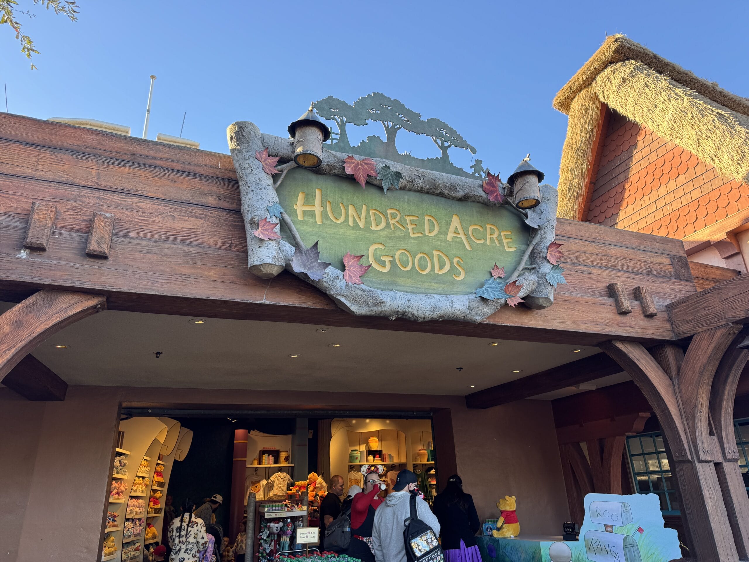Storefront with a sign reading "Hundred Acre Goods" evokes a hint of the Magic Kingdom. People are browsing inside the charming shop, nestled beneath wooden beams and thatched roofing, capturing an enchanting atmosphere worthy of any 2024 photo report.