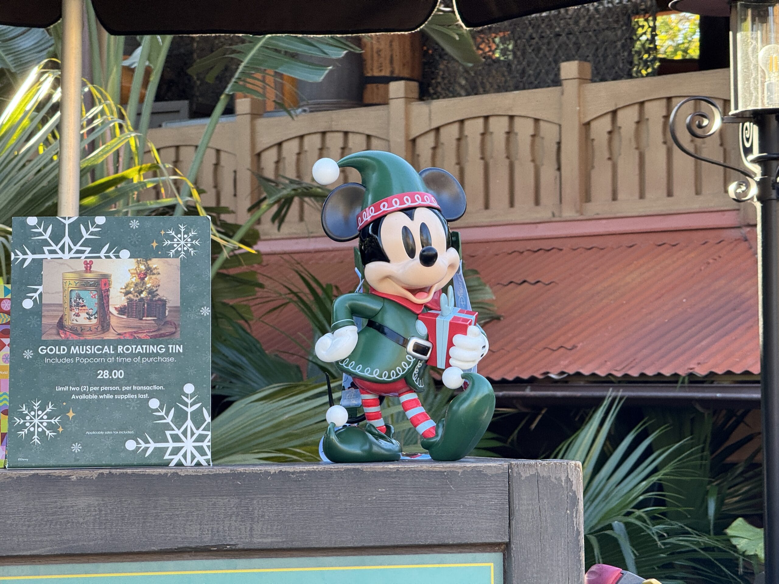 In a charming photo report from the Magic Kingdom, a festive Mickey Mouse figurine dressed in green, holding a present, stands beside a sign for a "Gold Musical Rotating Tin" priced at 28.00 in an enchanting outdoor setting. A delightful glimpse into the magic of 2024!