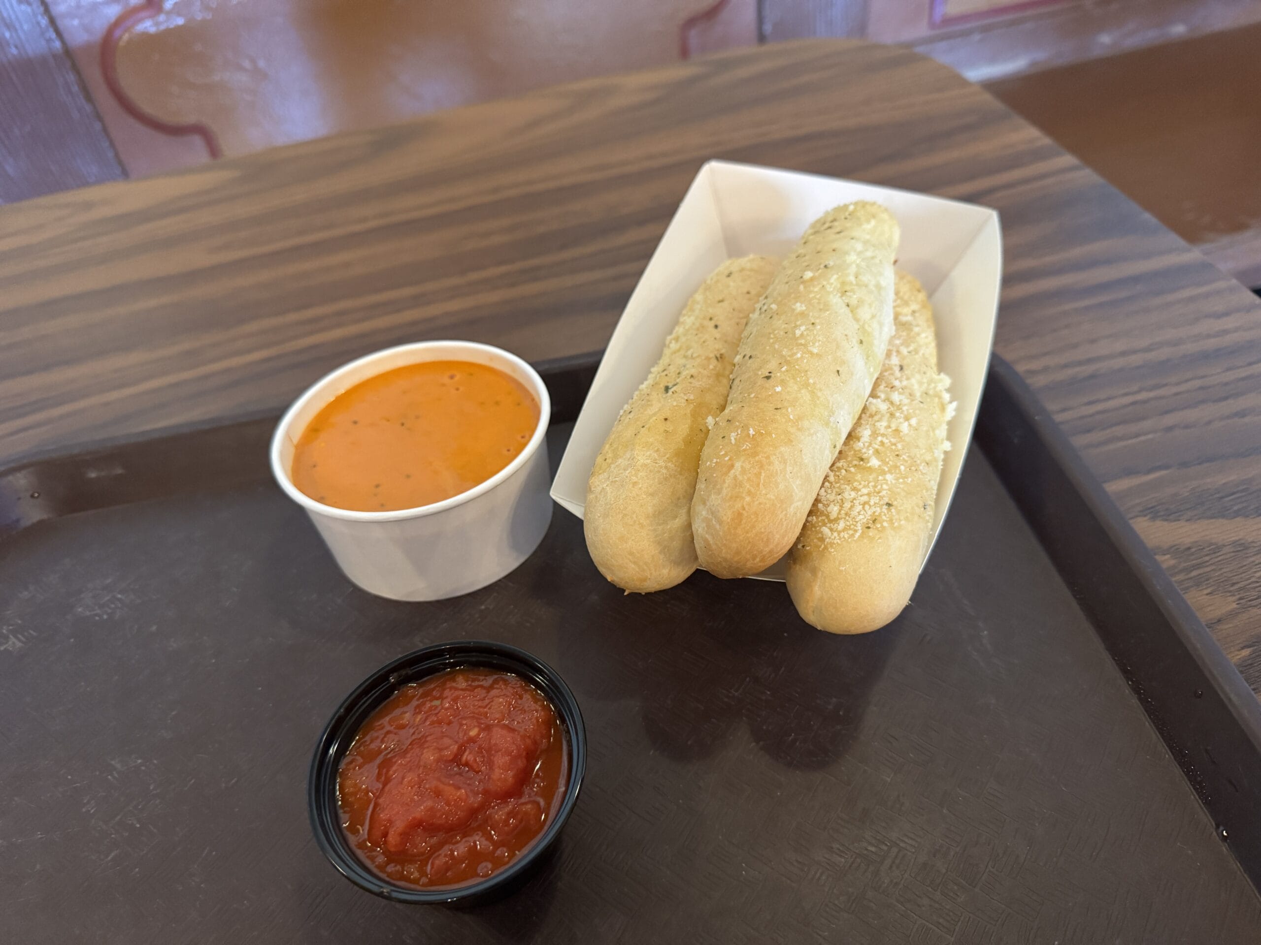 In a delightful photo report from Magic Kingdom, dated 12/7/2024, three seasoned breadsticks rest in a tray beside a cup of creamy tomato soup and a small container of red dipping sauce.
