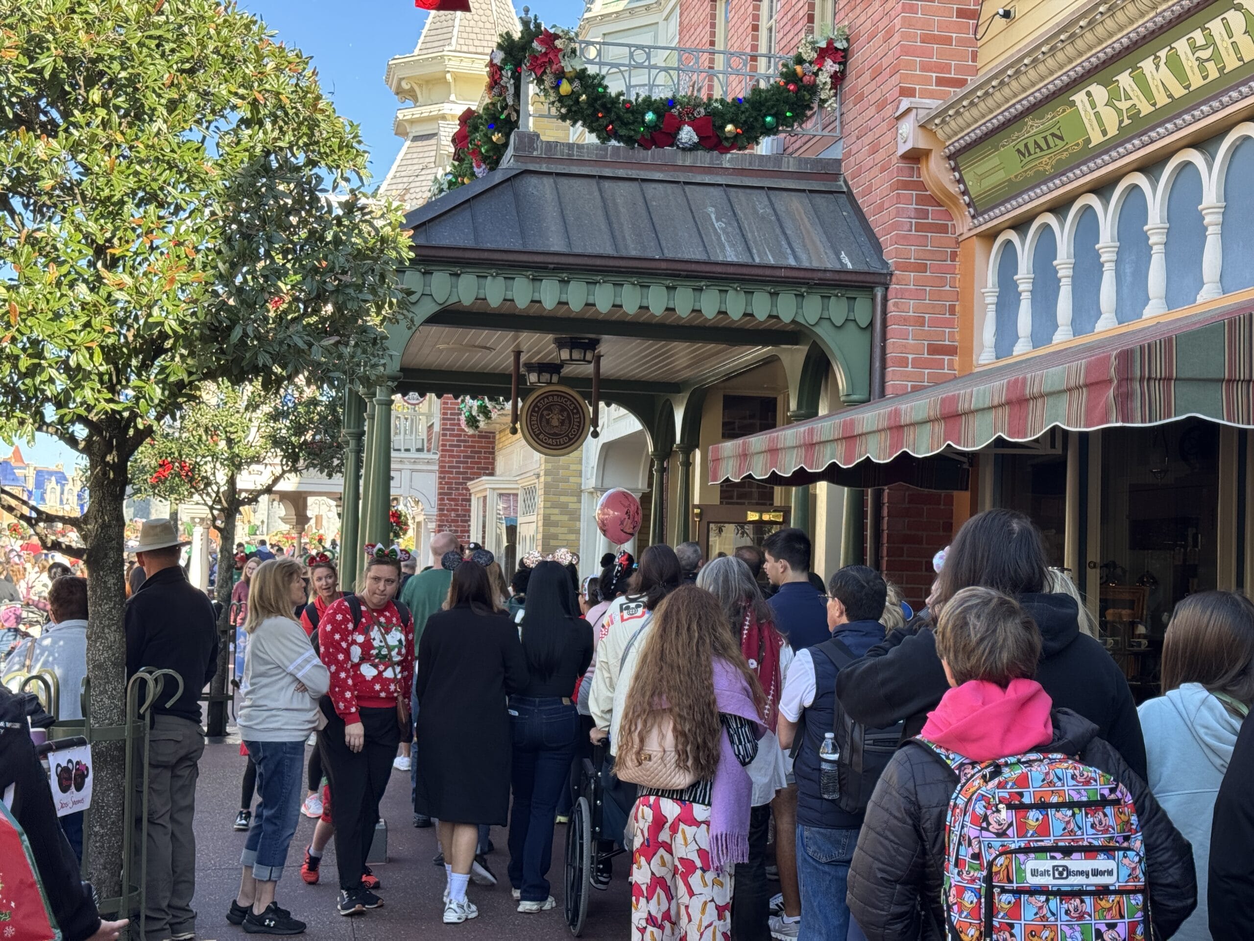 People wait in line outside a decorated bakery on a festive street with holiday garlands and wreaths, reminiscent of Magic Kingdom's charm. Some hold balloons, while one person sports a colorful backpack. This scene captures the delightful anticipation of 2024's holiday fervor.