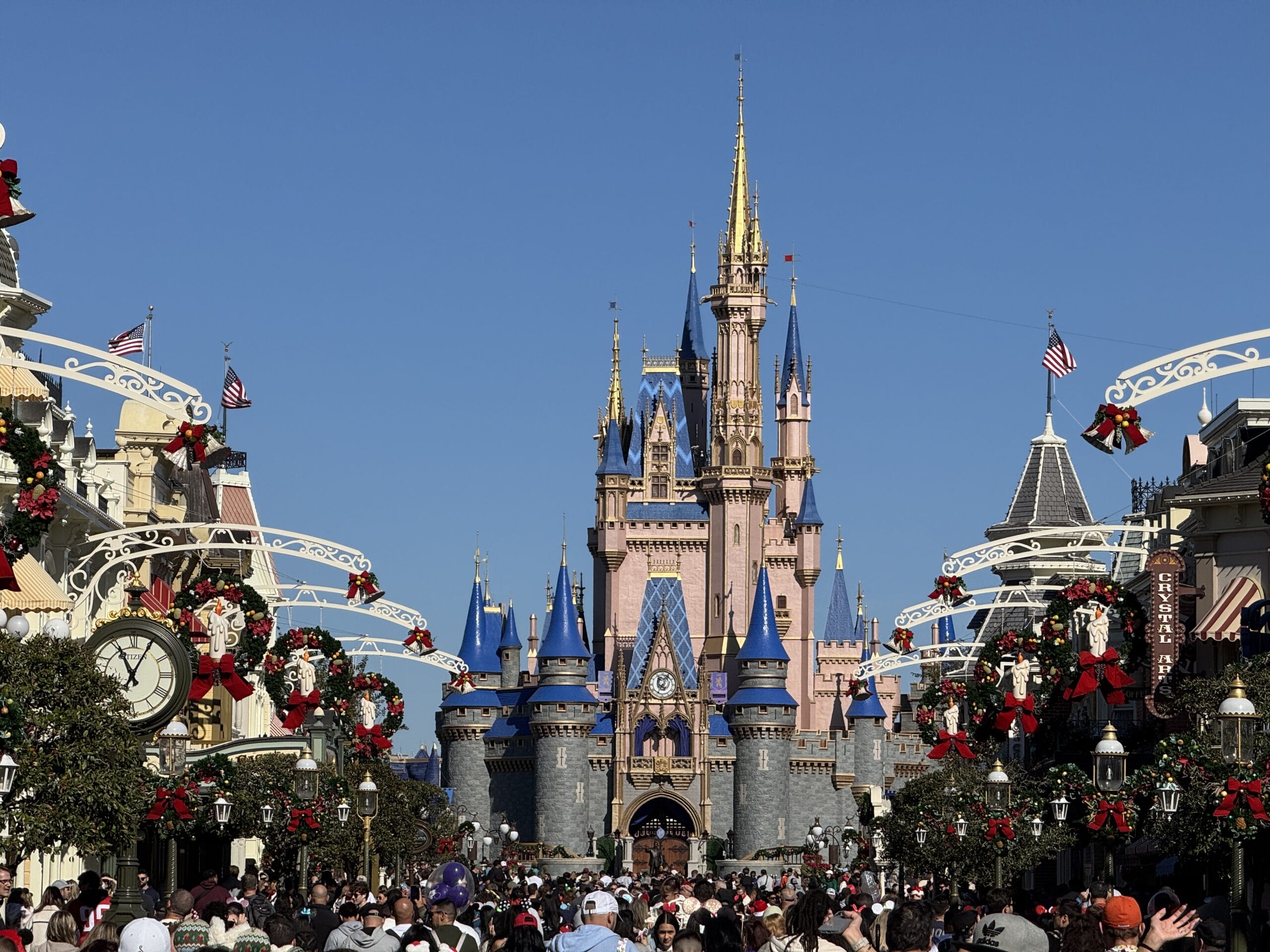 A crowd gathers at Magic Kingdom, facing a castle adorned with blue and gold spires. Holiday decorations and garlands line the street under a clear 2024 sky, capturing the enchantment in every moment for a captivating photo report.
