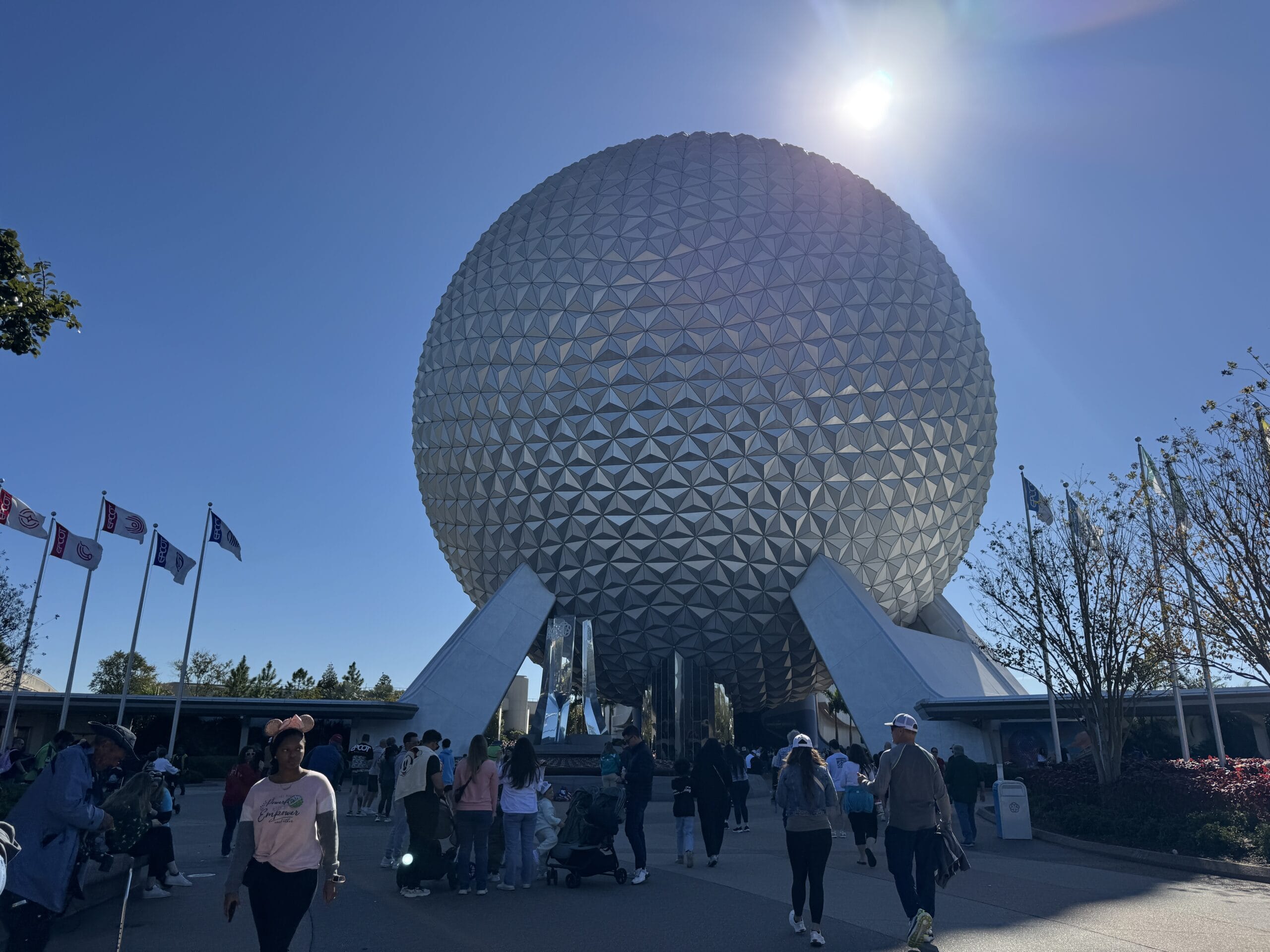 A large geodesic sphere stands against a clear blue sky with a sun flare in this 2024 photo report. People walk nearby, and flags are visible to the left, capturing the magical atmosphere reminiscent of the Magic Kingdom.