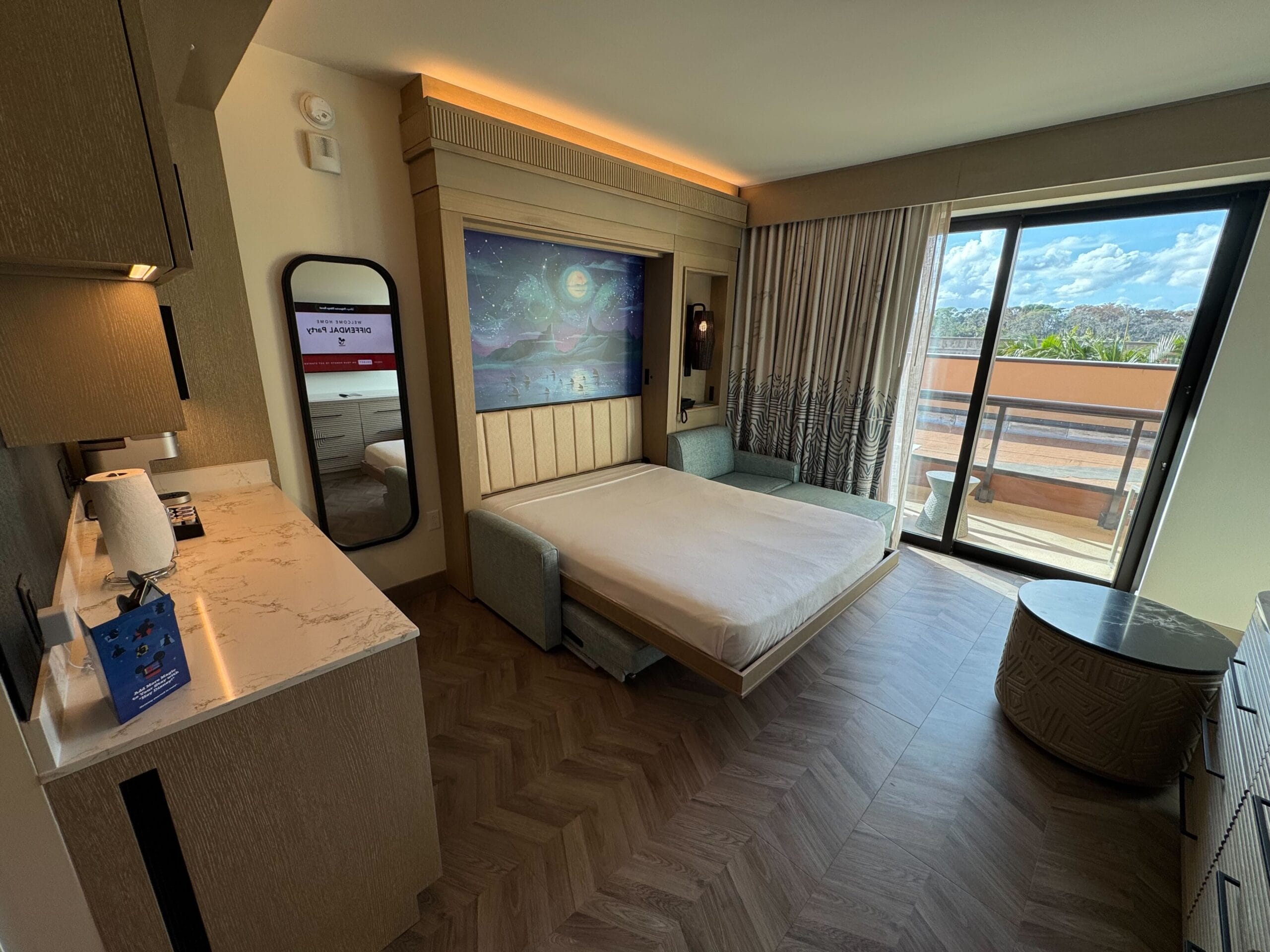 Hotel room with a pull-down bed, mirror, dresser, and balcony. A large wall painting decorates the space. The room has wood flooring and a view of the outdoors.
