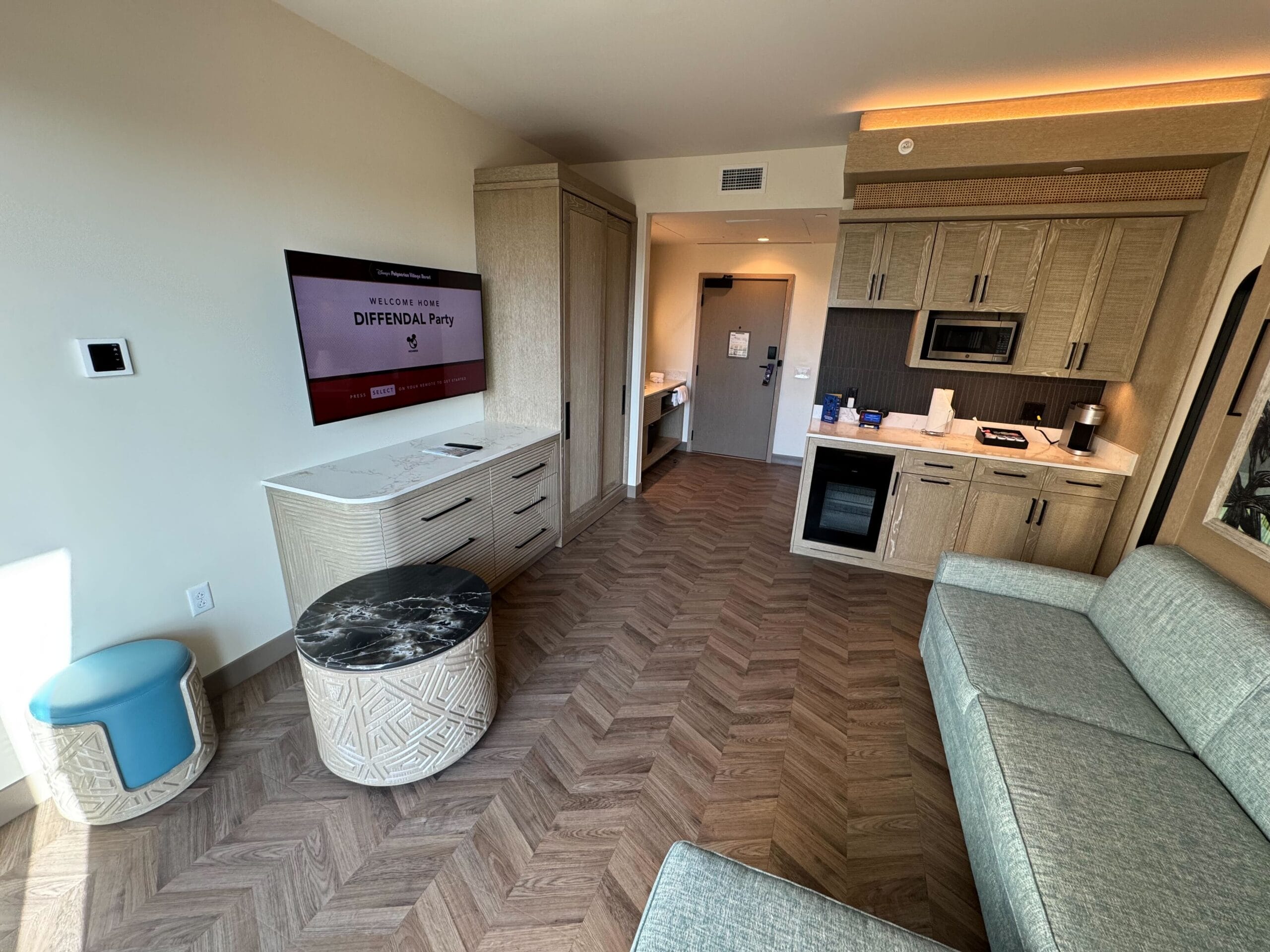 Hotel room with wooden flooring, a kitchenette with appliances, a wall-mounted TV displaying "DIFFENDAL Party," a sofa, and a small round table with a blue stool. Door visible in the background.
