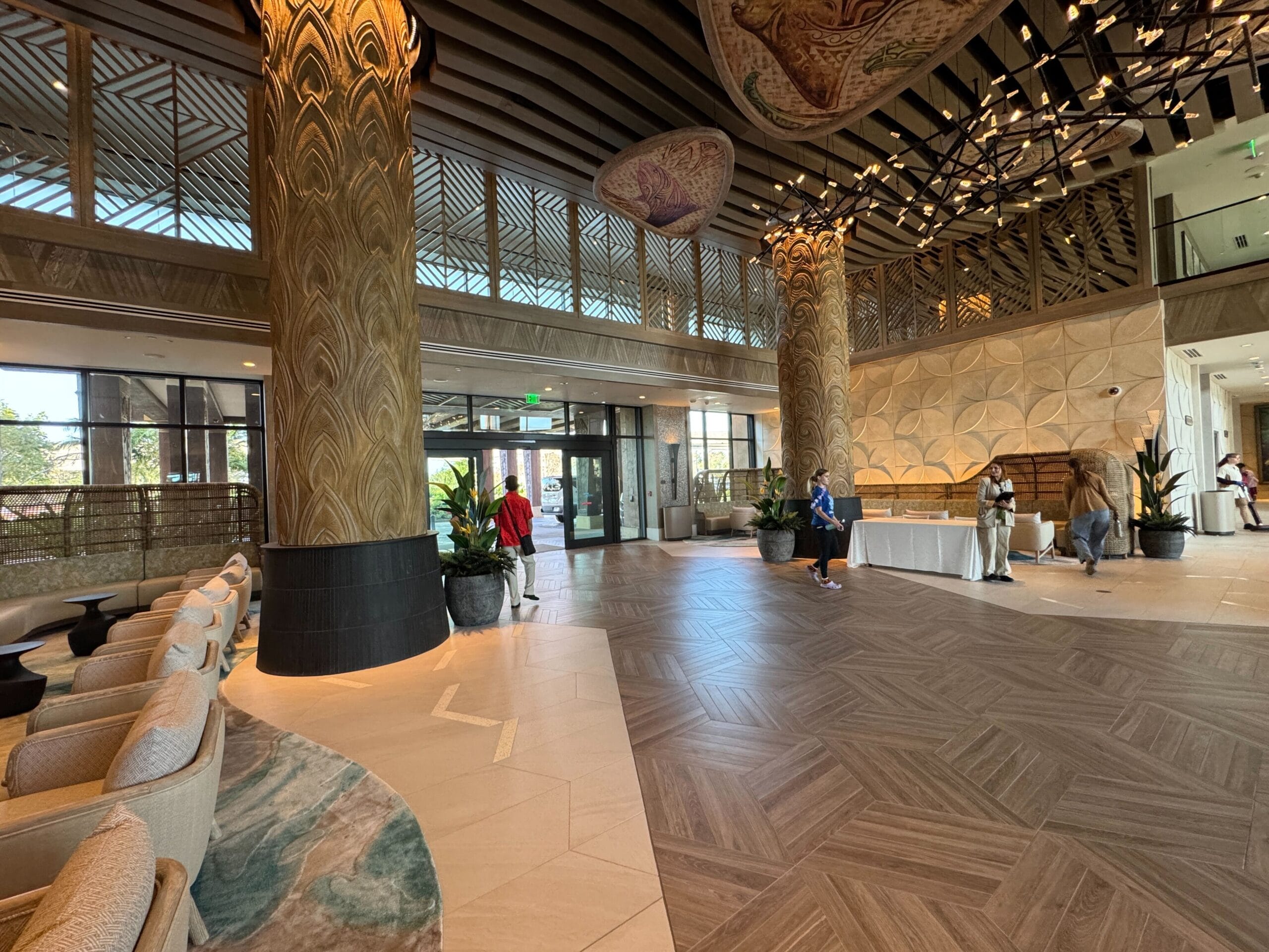 A spacious hotel lobby with ornate columns, seating areas, and large windows. Guests walk through, with a reception desk on the right. Ceiling features decorative patterns and lighting.
