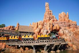 A yellow train races on a wooden track through red rocks on Big Thunder Mountain Railroad under a clear blue sky.