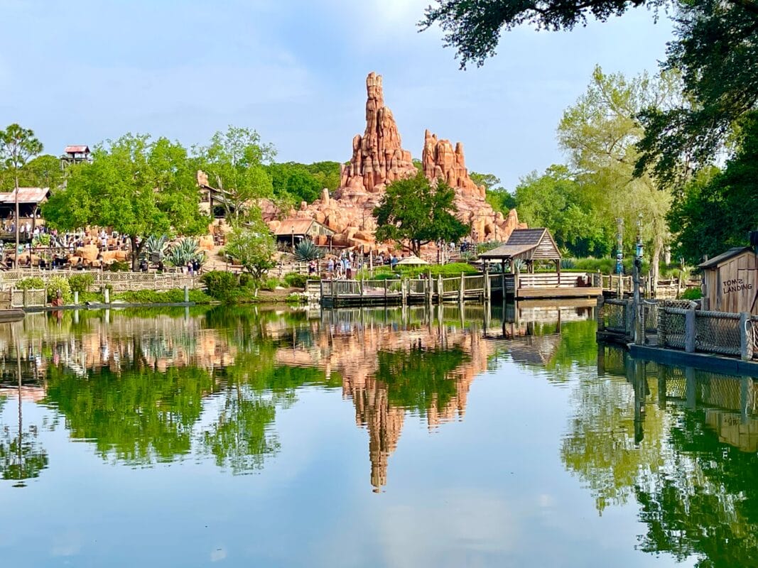 Big Thunder Mountain Railroad reflected in a calm lake, surrounded by greenery and wooden structures under a clear sky.