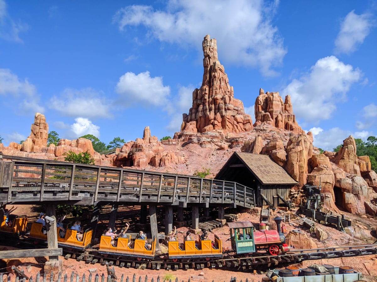 A Big Thunder Mountain Railroad train races through a rocky, mountainous landscape under a blue sky with scattered clouds.
