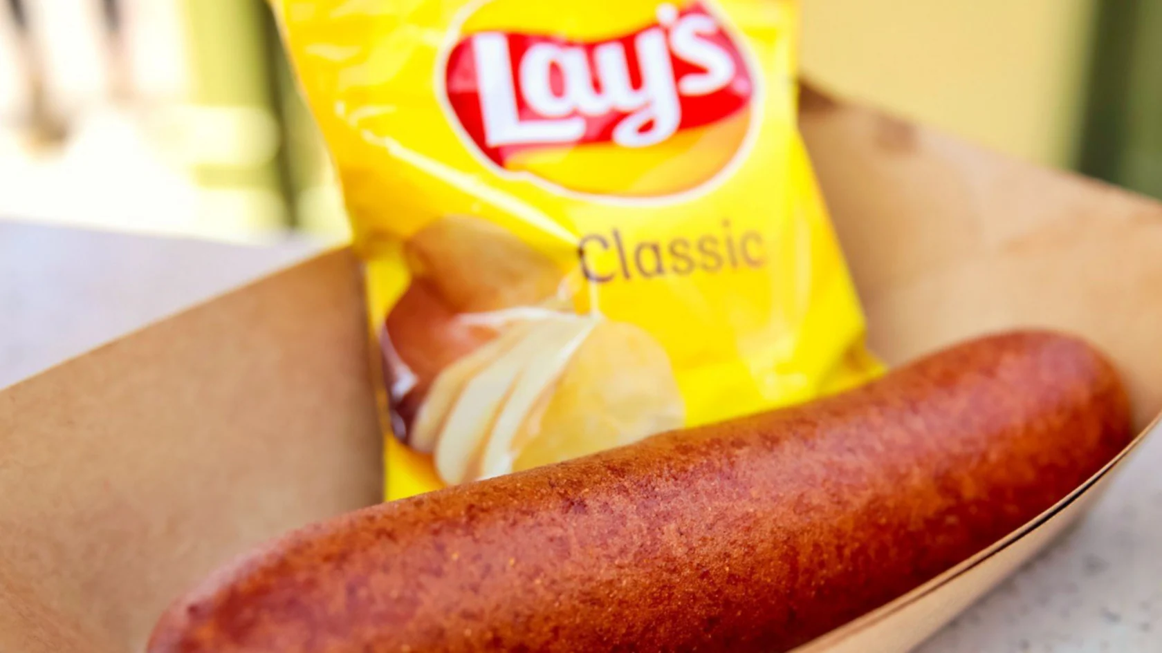 A Blue Ribbon Corn Dog in a tray with a bag of Lay's Classic potato chips in the background.