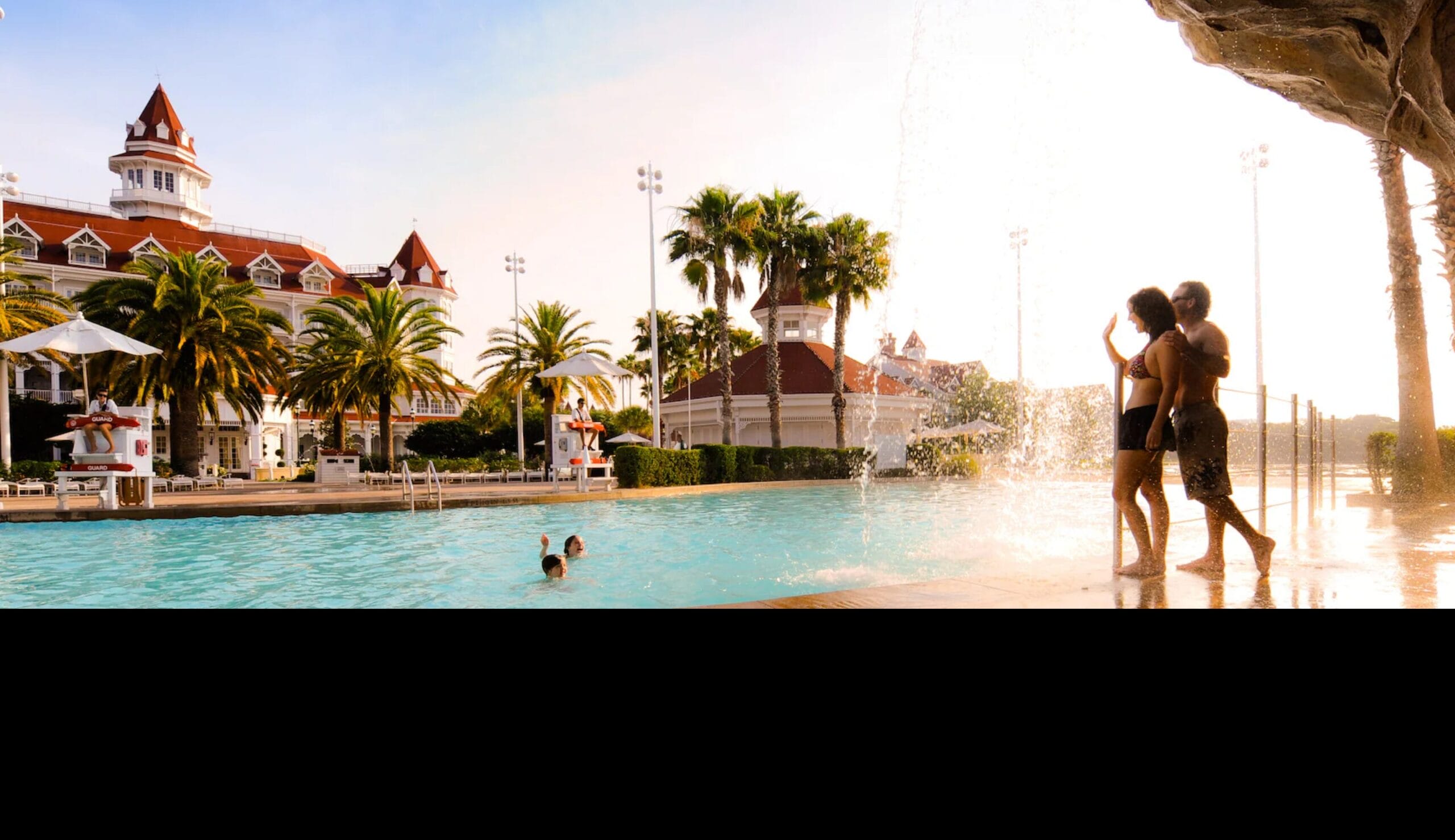 A couple stands by a resort pool bar as children swim, with palm trees and a grand hotel in the background.