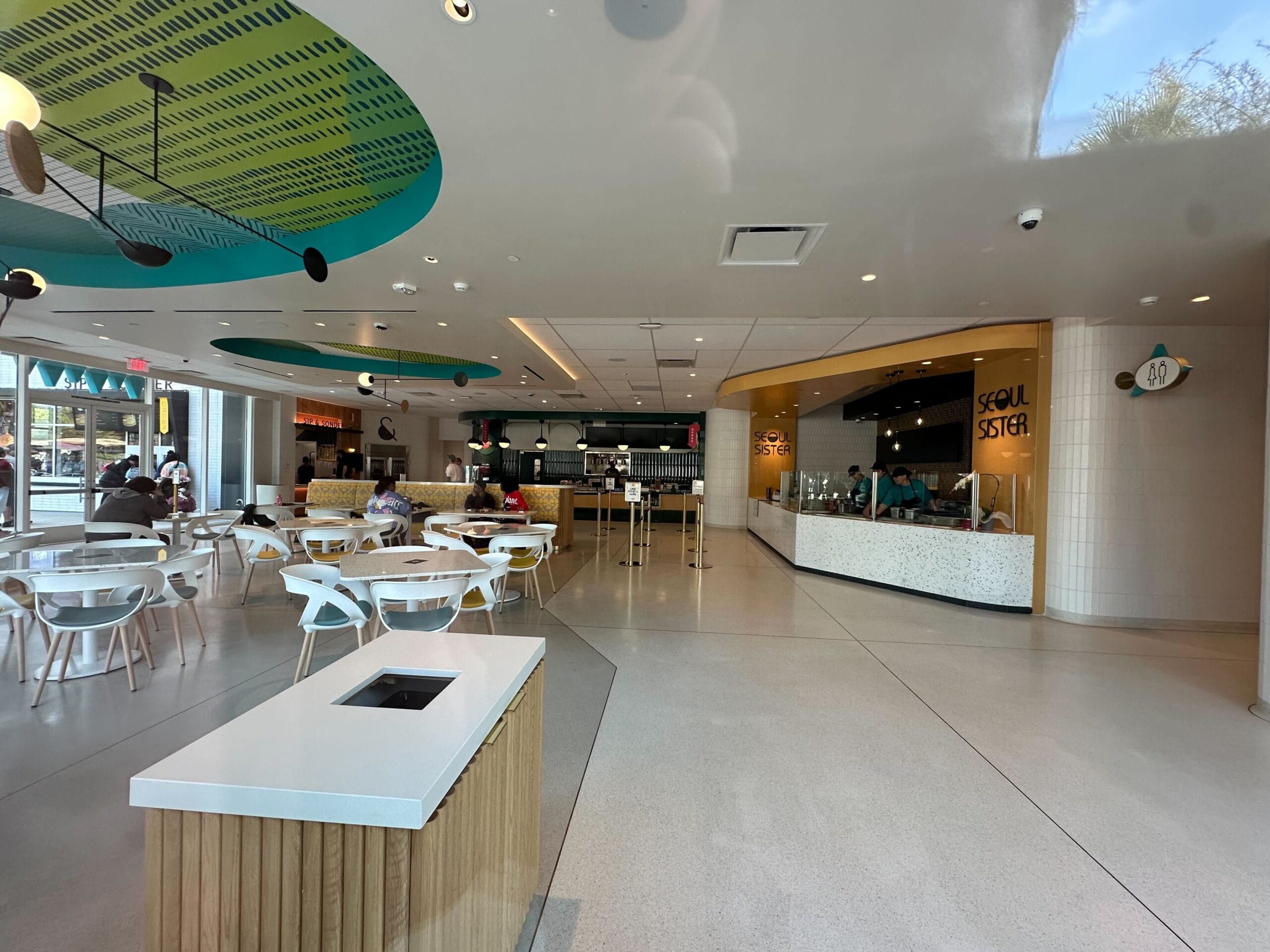 Explore the modern cafeteria at Parkside Market, featuring white tables and chairs with several people seated. On the right, the food counter labeled "Seoul Sister" adds charm to this bright, open space with teal ceiling accents. It's a dining experience reminiscent of Disney Springs magic.