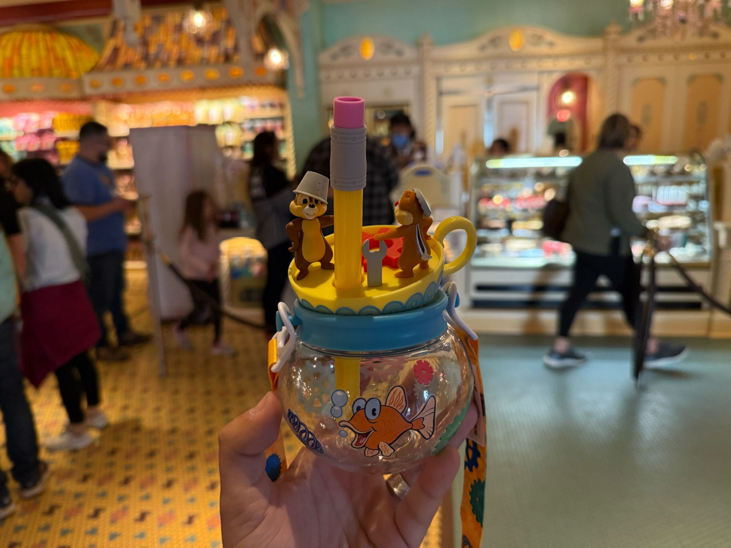 A hand holds a decorated mug with cartoon characters and a straw. The background shows a busy indoor setting with people and a display case.