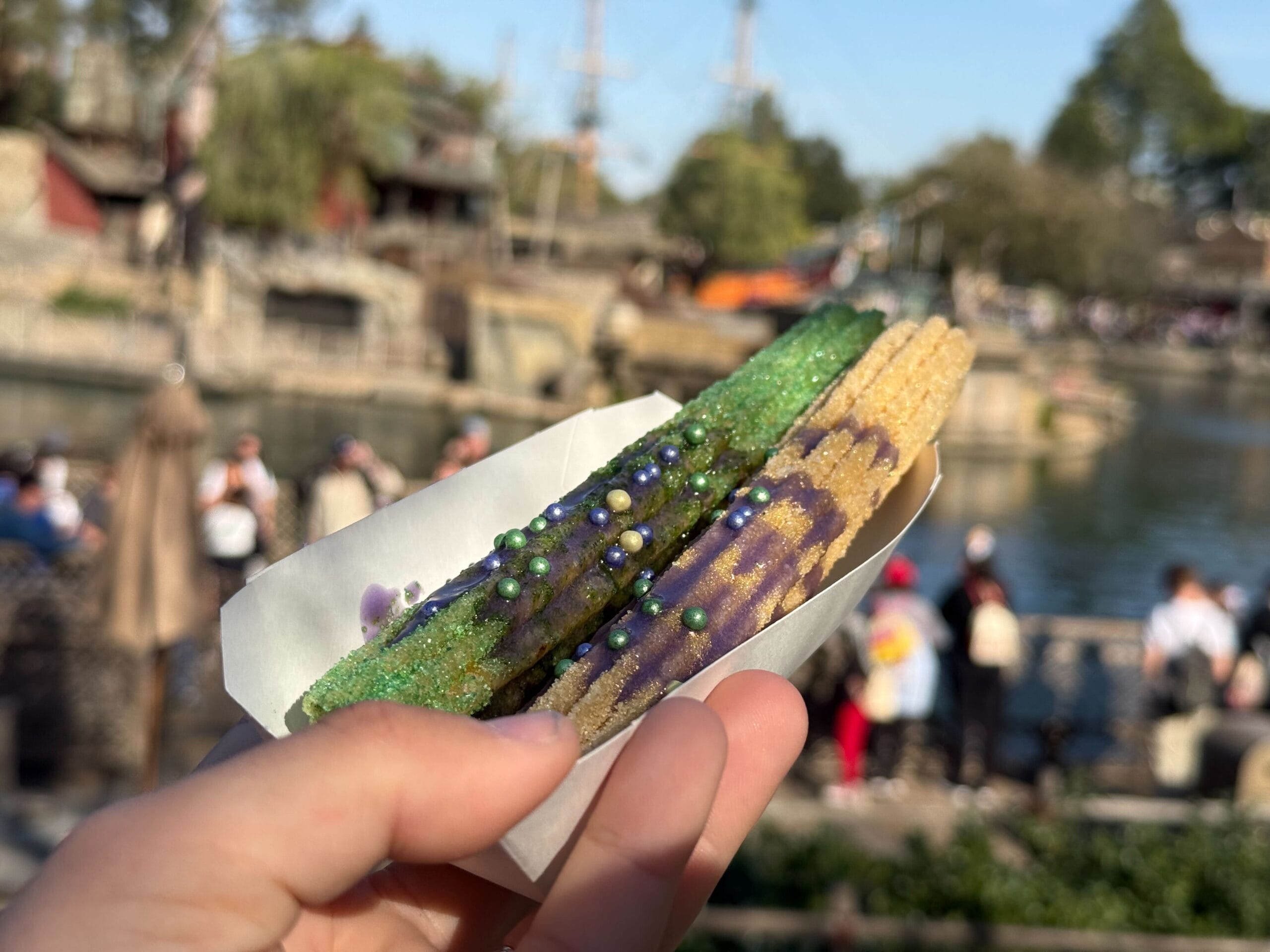 A hand holds two churros covered in colorful sugar and edible pearls, with a scenic riverside background and people in the distance.