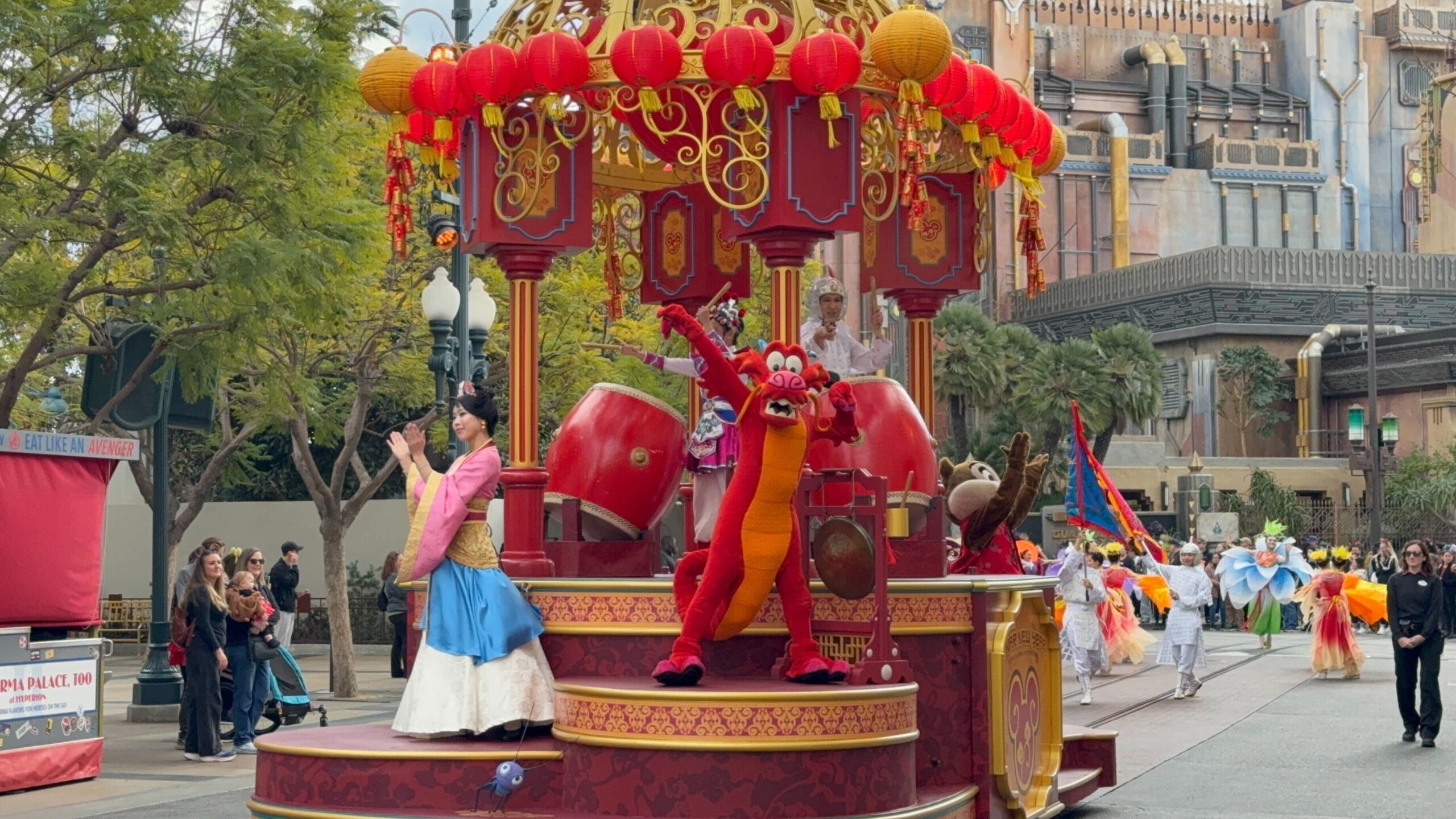 The parade float with a red and gold theme channels Mulan's Lunar New Year Procession, featuring performers in vibrant costumes and a dragon character, all set against an urban backdrop filled with trees and intrigued onlookers.