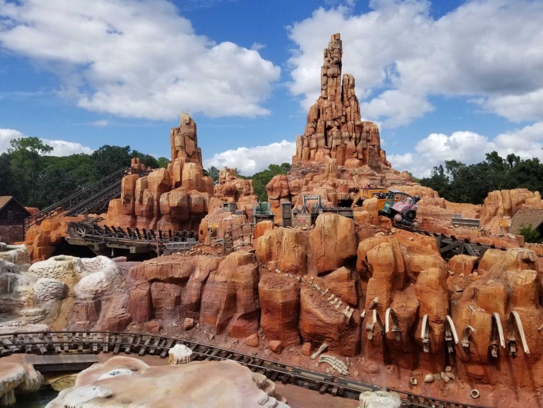 A train roller coaster winds through red rocks and clouds on Big Thunder Mountain Railroad at an amusement park.