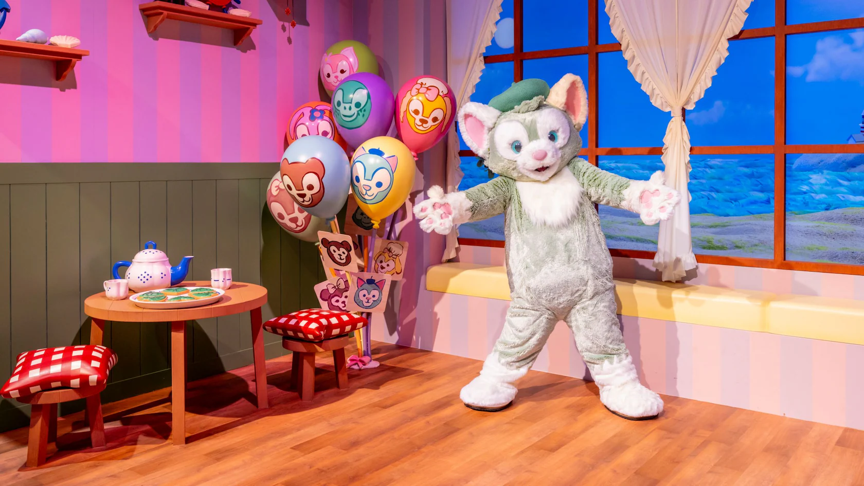 In a colorful Friends Play House, a person in a cat costume poses amidst balloons. A small table with a tea set and cupcakes invites whimsy, while blue curtains frame the scene, evoking the playful charm of Duffy and Gelatoni's world.