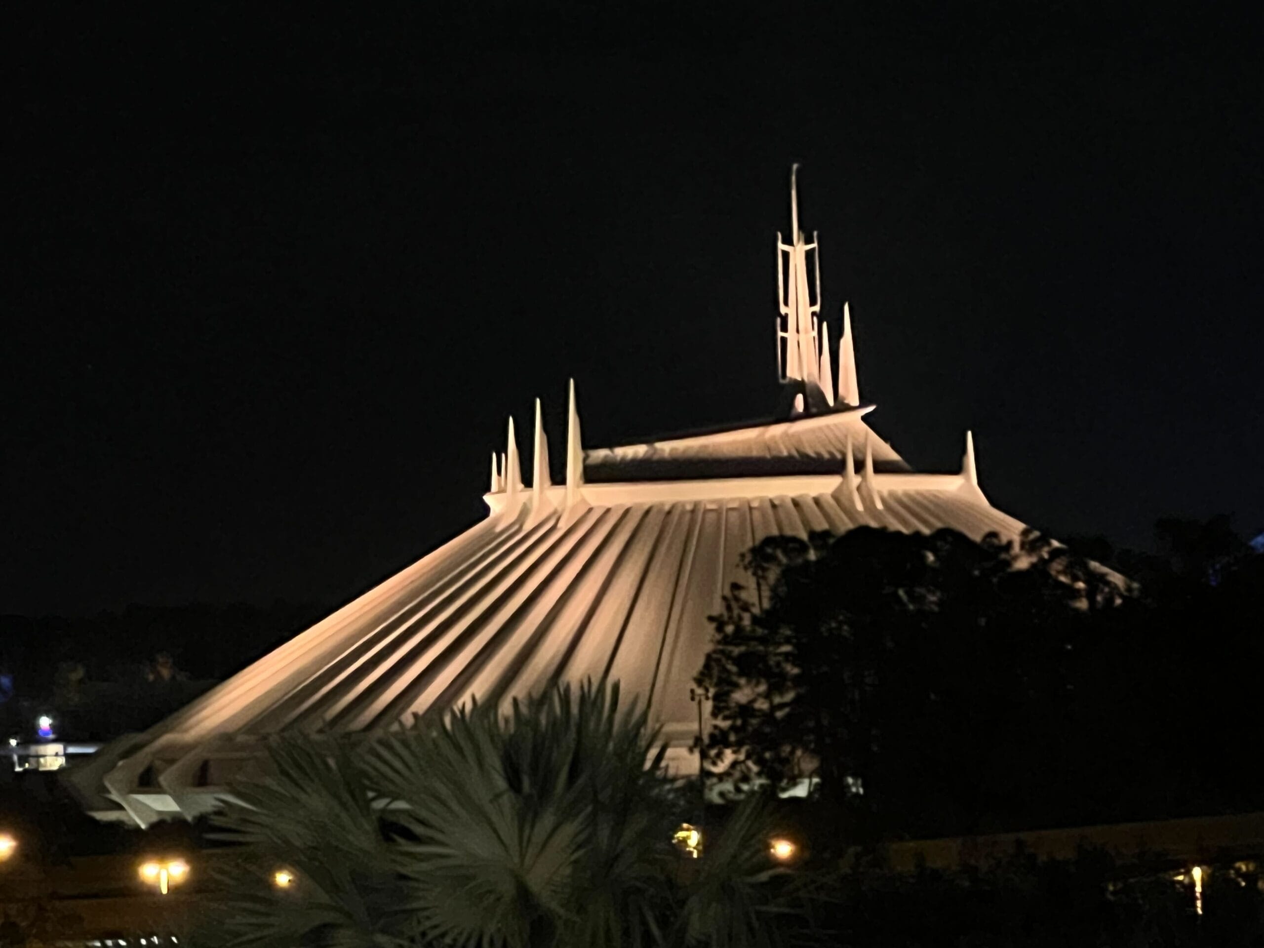 Night view of a futuristic, illuminated building with pointed spires and a sloped roof, surrounded by trees.