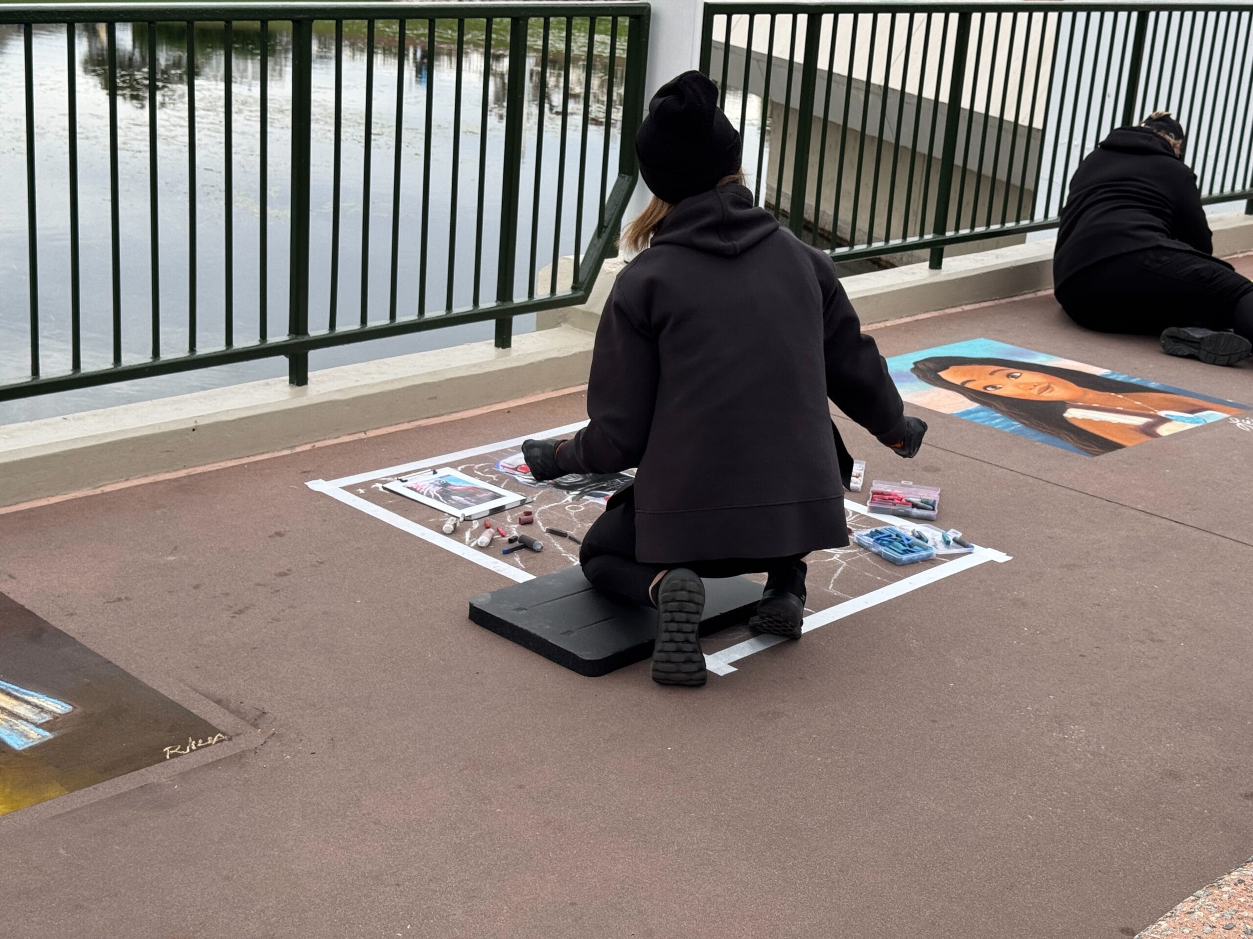 At the Festival of the Arts, a person in black clothing skillfully creates vibrant chalk art on the pavement by the water's edge, just steps from a bustling EPCOT crowd. Nearby, another artist works diligently, adding to the colorful display.