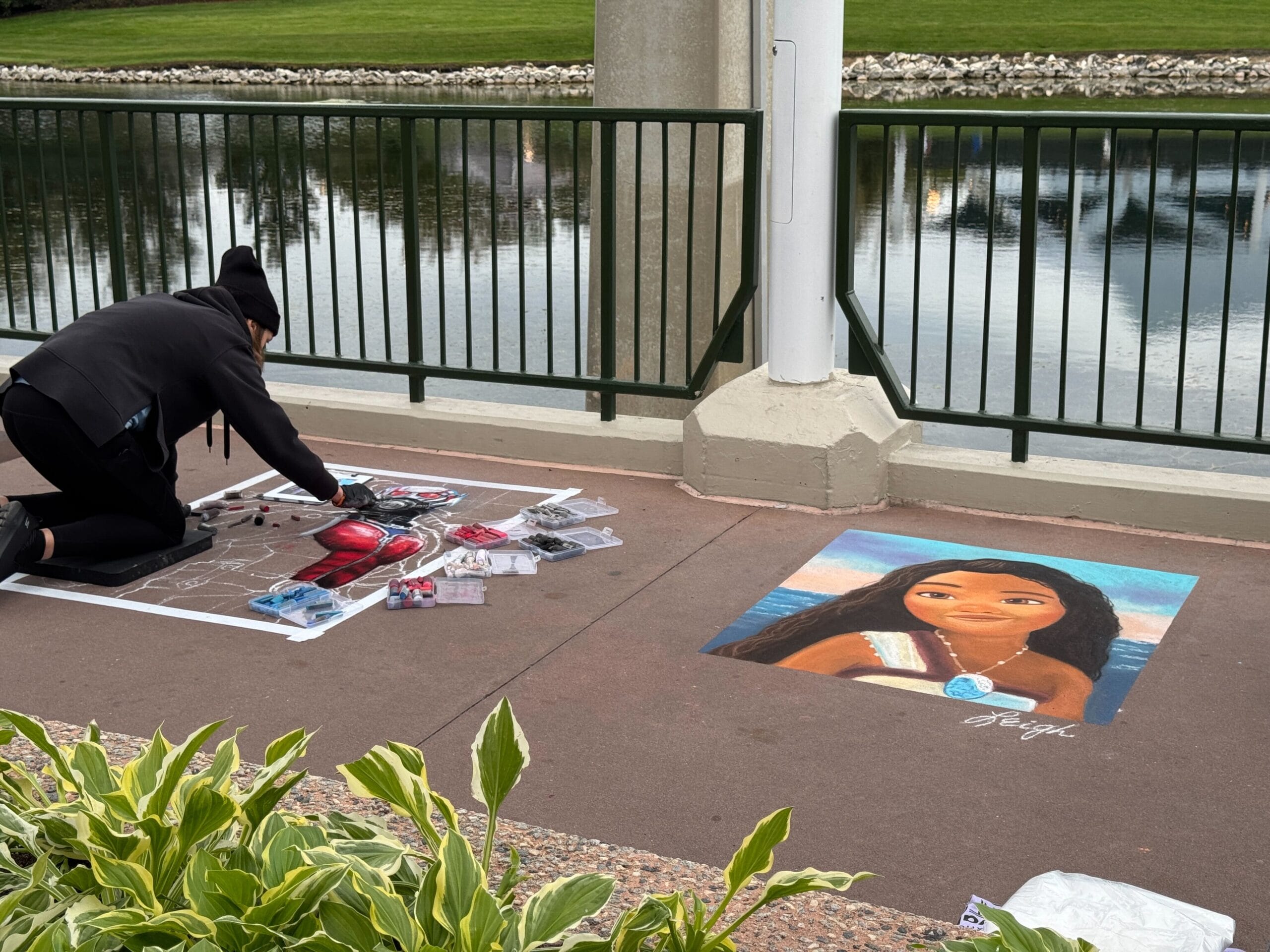 An artist brings the riverside path to life with chalk art, illustrating a girl with long hair. Supplies are scattered around, reminiscent of the vibrant energy found at EPCOT's Festival of the Arts—a perfect setting for creativity and inspiration.