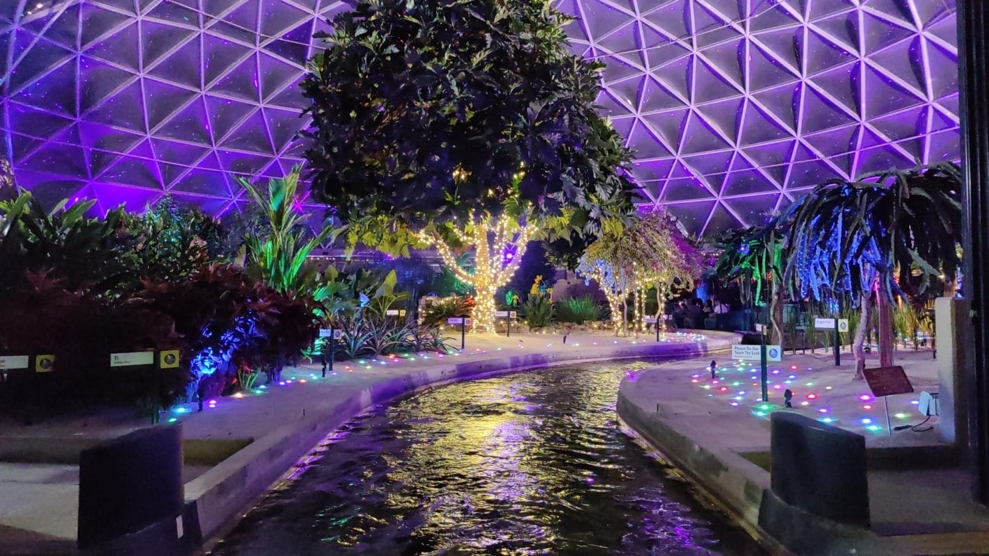 Indoor garden with colorful lights and winding waterway inspired by Living with the Land at Epcot under a purple-lit dome.