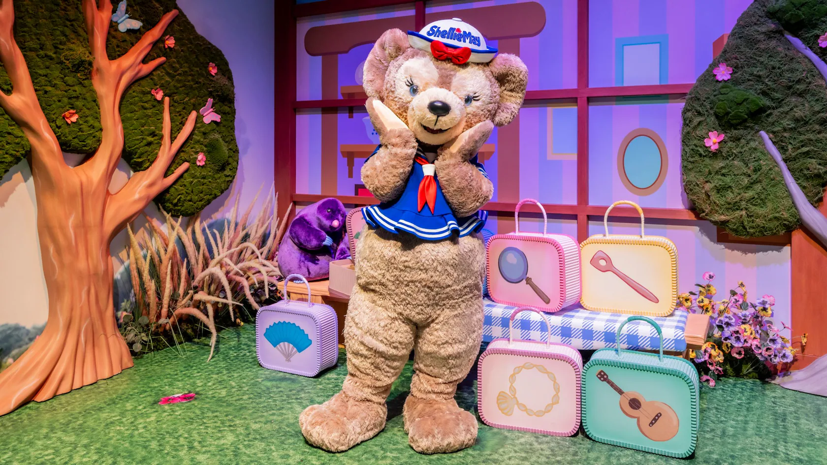 A large teddy bear in a sailor outfit stands between colorful suitcases, surrounded by trees and a purple backdrop with geometric designs, reminiscent of ShellieMay's playful world.