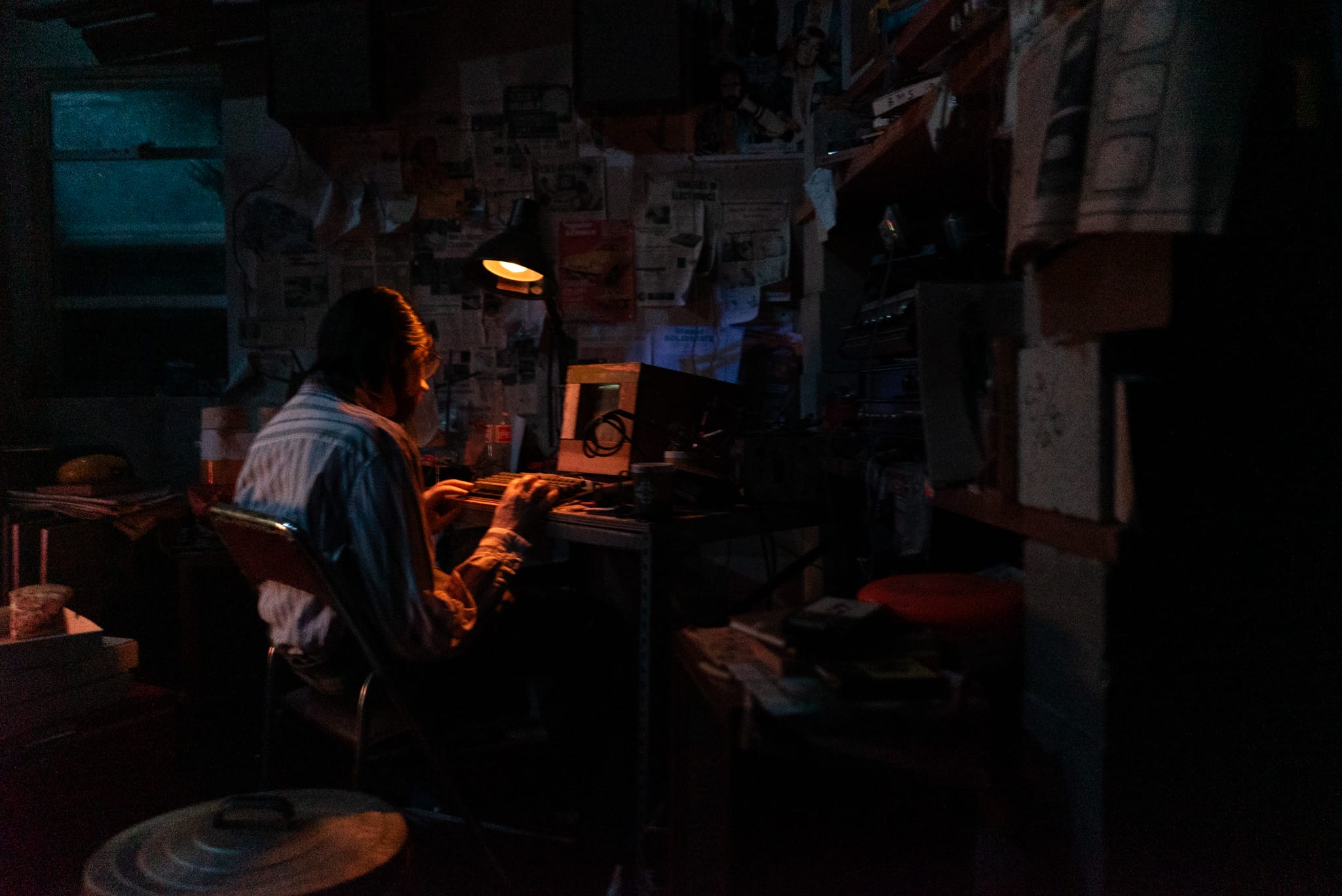 A person sits alone at a cluttered desk, typing in a dim room filled with papers, like the captain of Spaceship Earth.