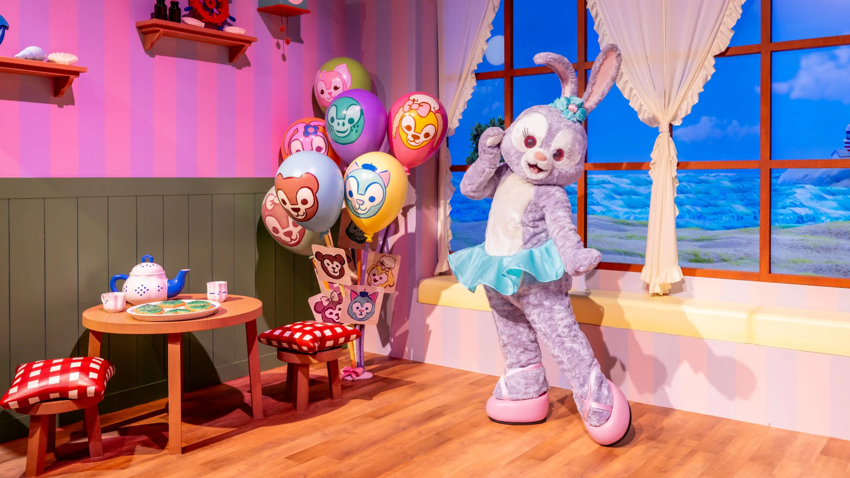 In the playful setting of a colorful room resembling a Play House, StellaLou, in her blue dress and bunny ears, stands amidst balloons adorned with cartoon faces. Nearby, a small table with a tea set awaits under the gaze of Duffy and Friends through a scenic window view.