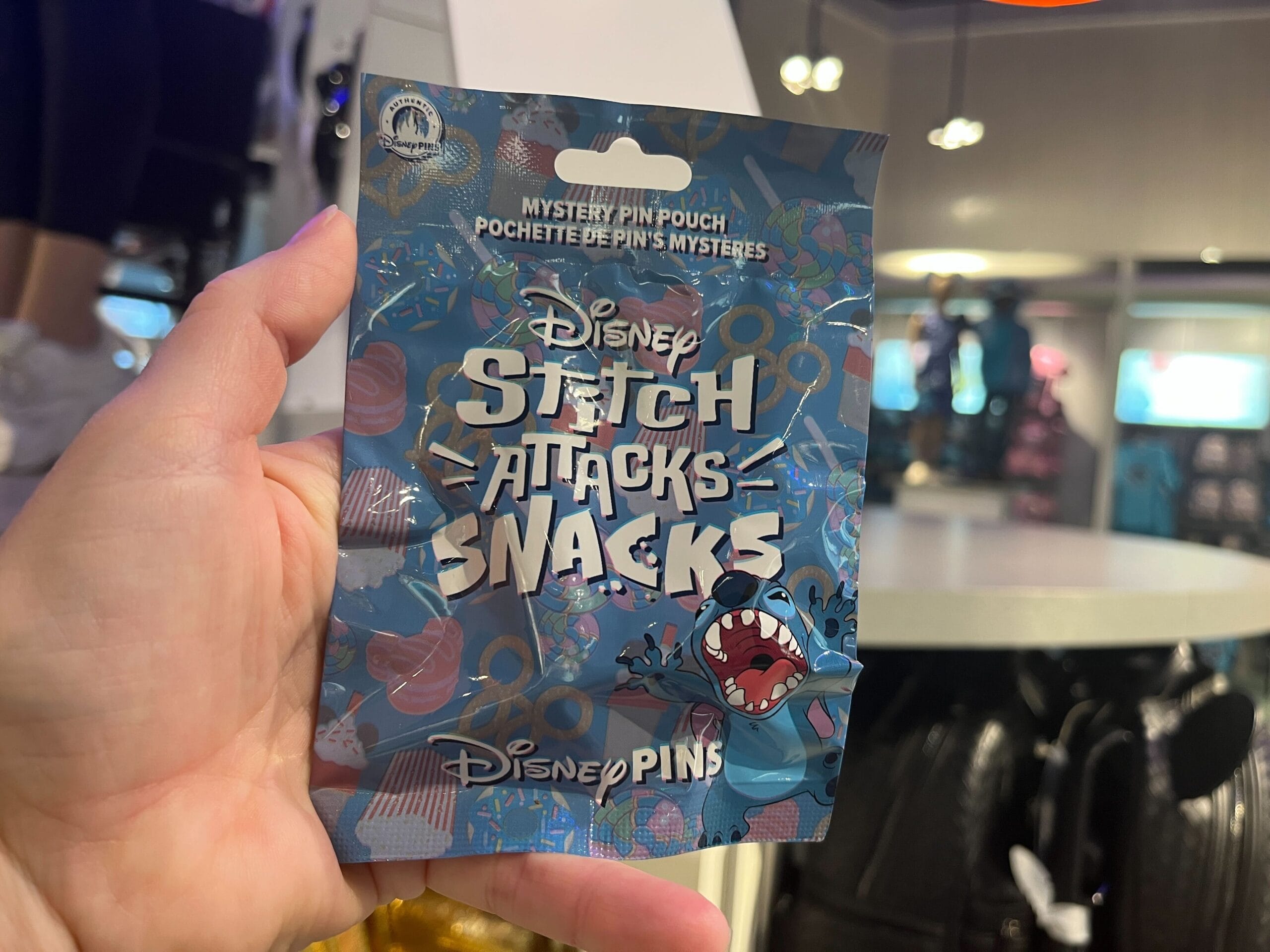 A hand holds a Disney Stitch Attacks Snacks mystery pin pouch, showcasing colorful packaging adorned with playful Stitch imagery, inside a bustling store.