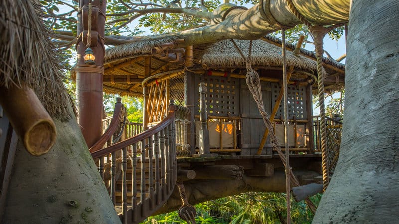 A wooden Swiss Family Treehouse with a thatched roof, nestled among large branches and lush greenery.