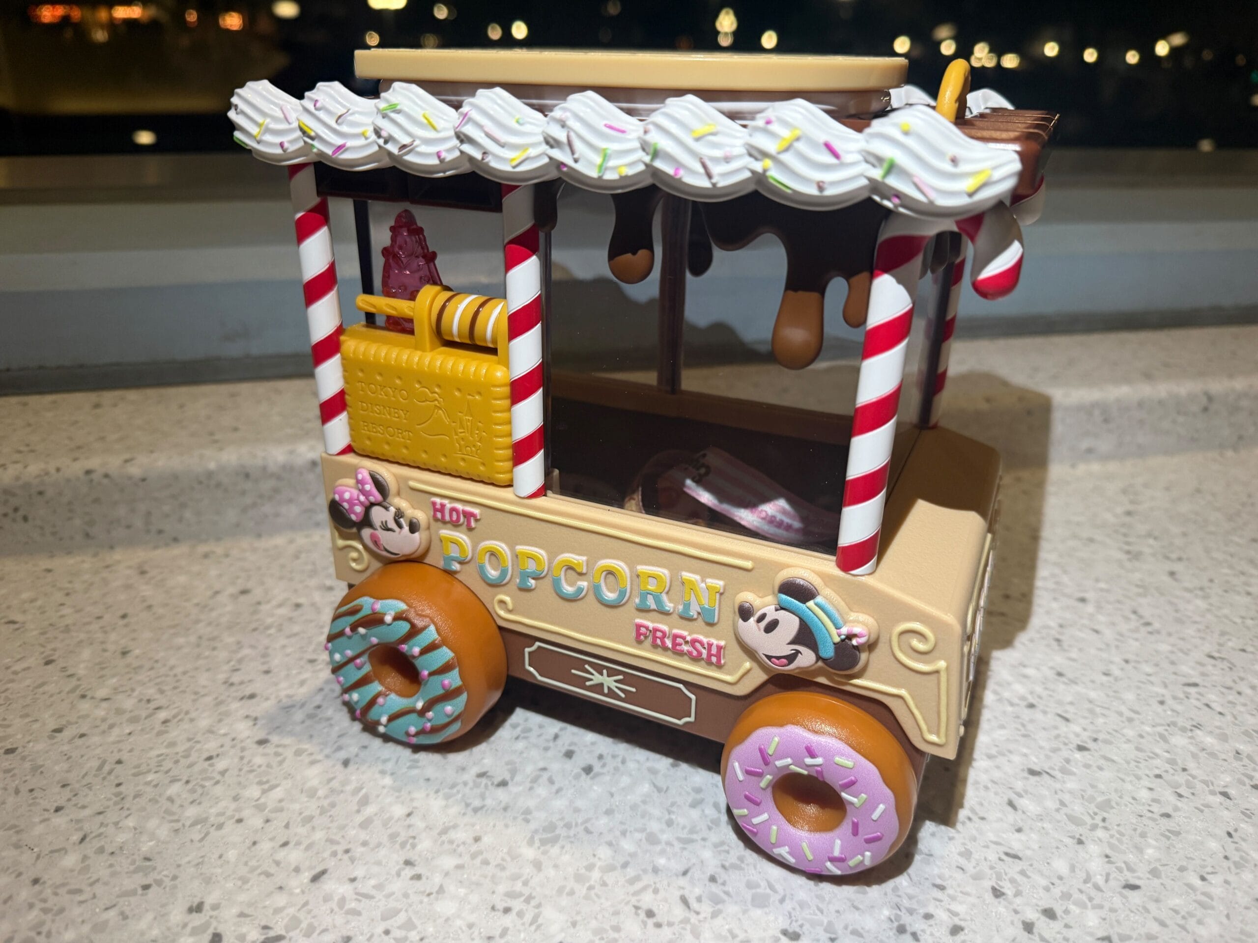 A toy popcorn cart decorated with candy-themed elements, including donut wheels and a striped canopy, is placed on a countertop.