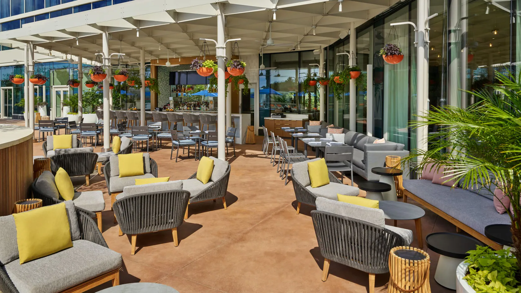 Outdoor restaurant patio with modern gray chairs, yellow cushions, small tables, and hanging plants by large glass windows.