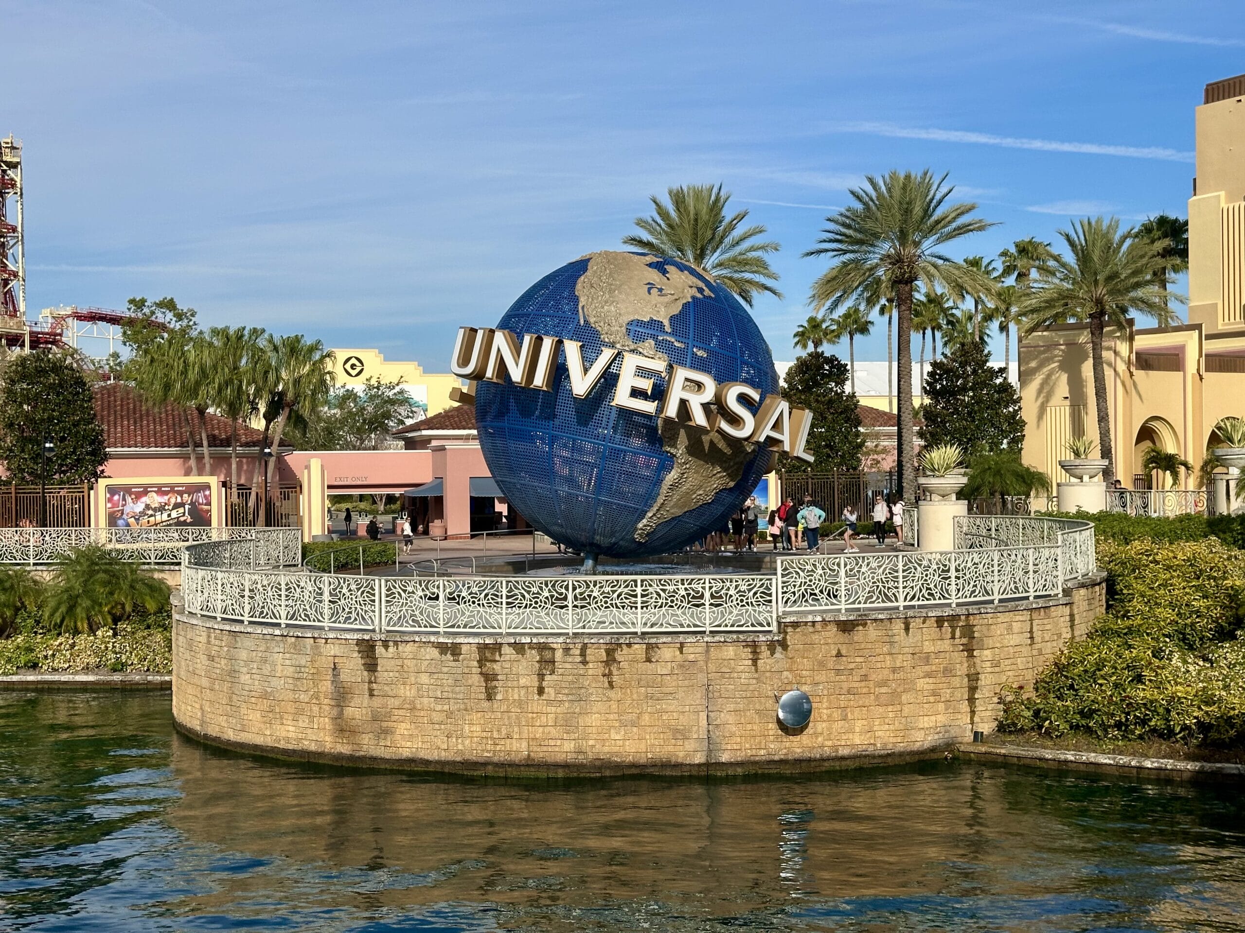 Giant blue globe with "Universal" text, surrounded by palm trees and buildings, in front of a water feature at Universal Orlando, showcasing vibrant Mardi Gras decor.