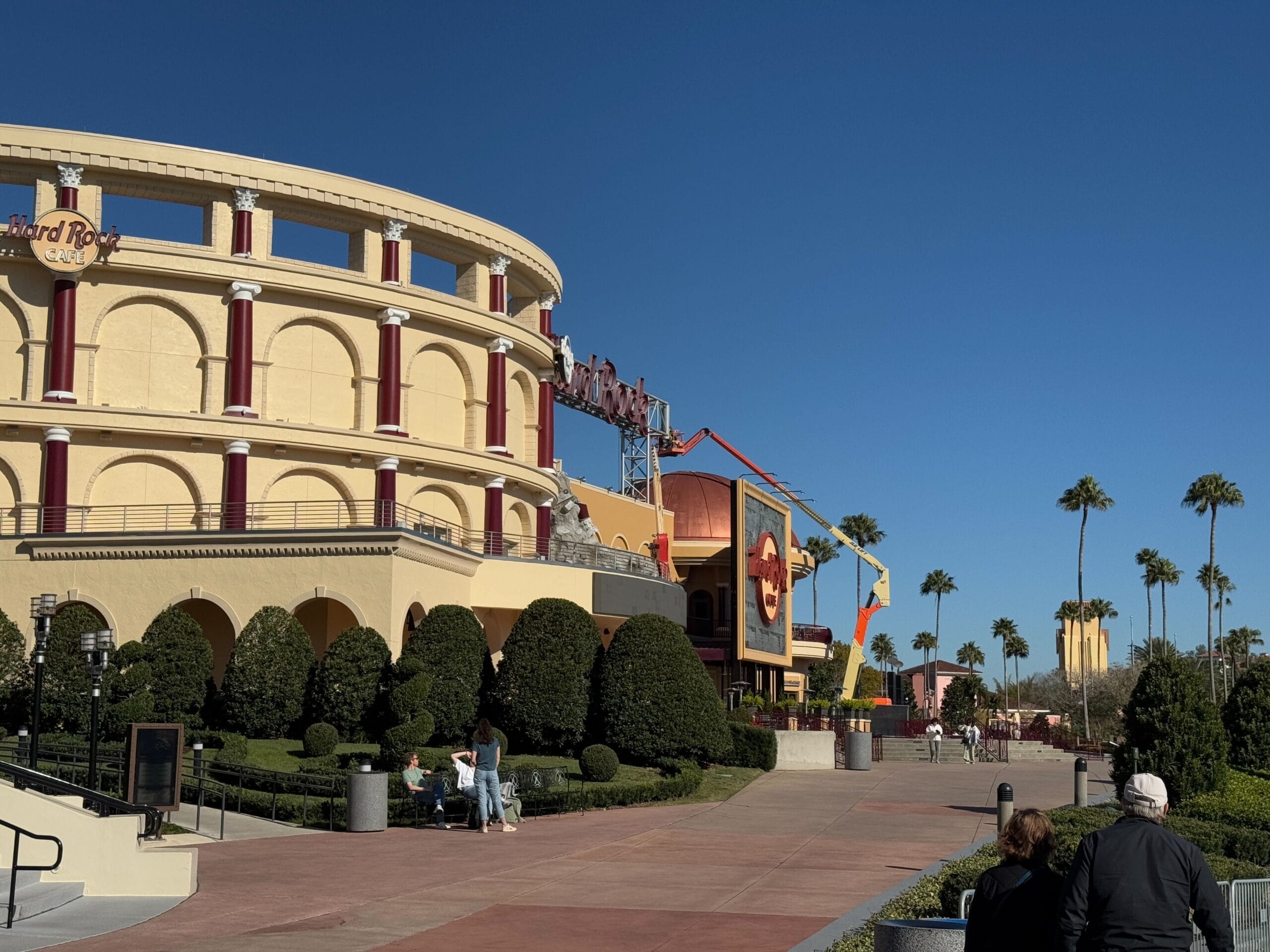 Wide walkway lined with trees leads to a beige, round building with red accents and a guitar sign, reminiscent of the iconic Hard Rock Cafe at Universal Orlando CityWalk. People stroll through the sunny area, admiring the recent roof repainting that adds a fresh touch.