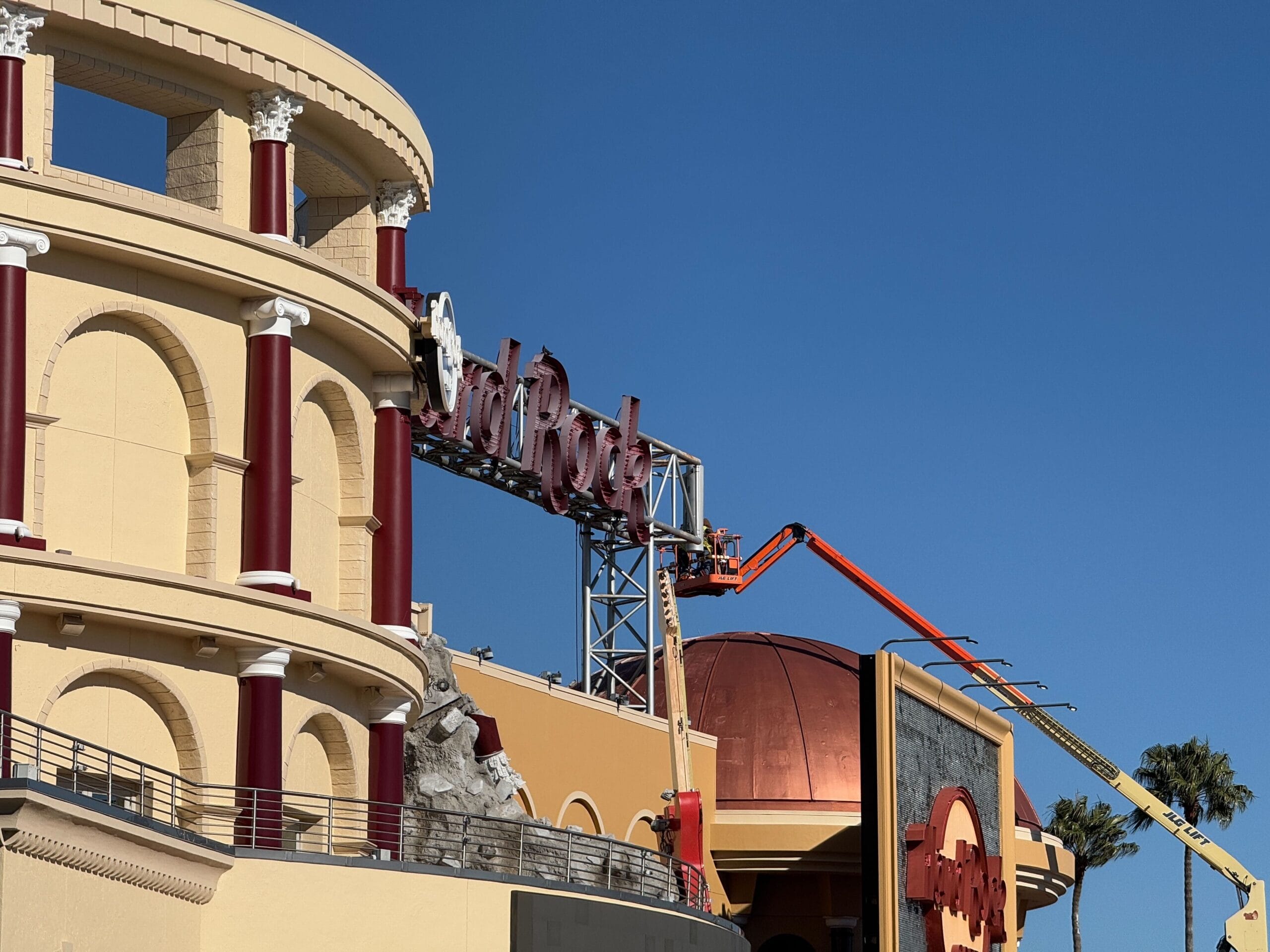 Workers in cherry pickers dismantle the "Hard Rock Cafe" sign on a beige and red building at Universal Orlando CityWalk under a clear blue sky.