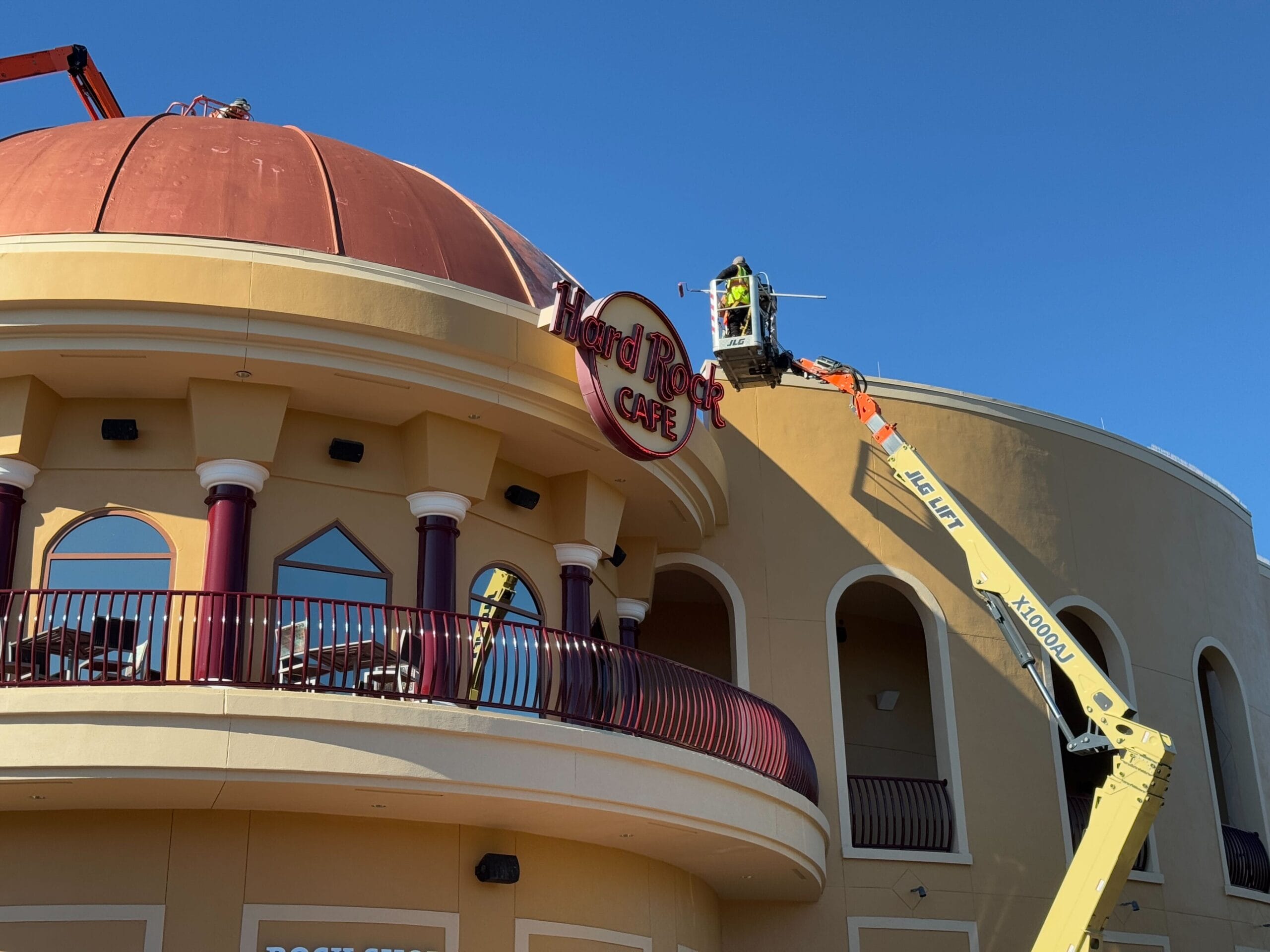 Workers on cranes installing a "Hard Rock Cafe" sign on a round building with a newly repainted roof under the clear blue sky at Universal Orlando CityWalk.