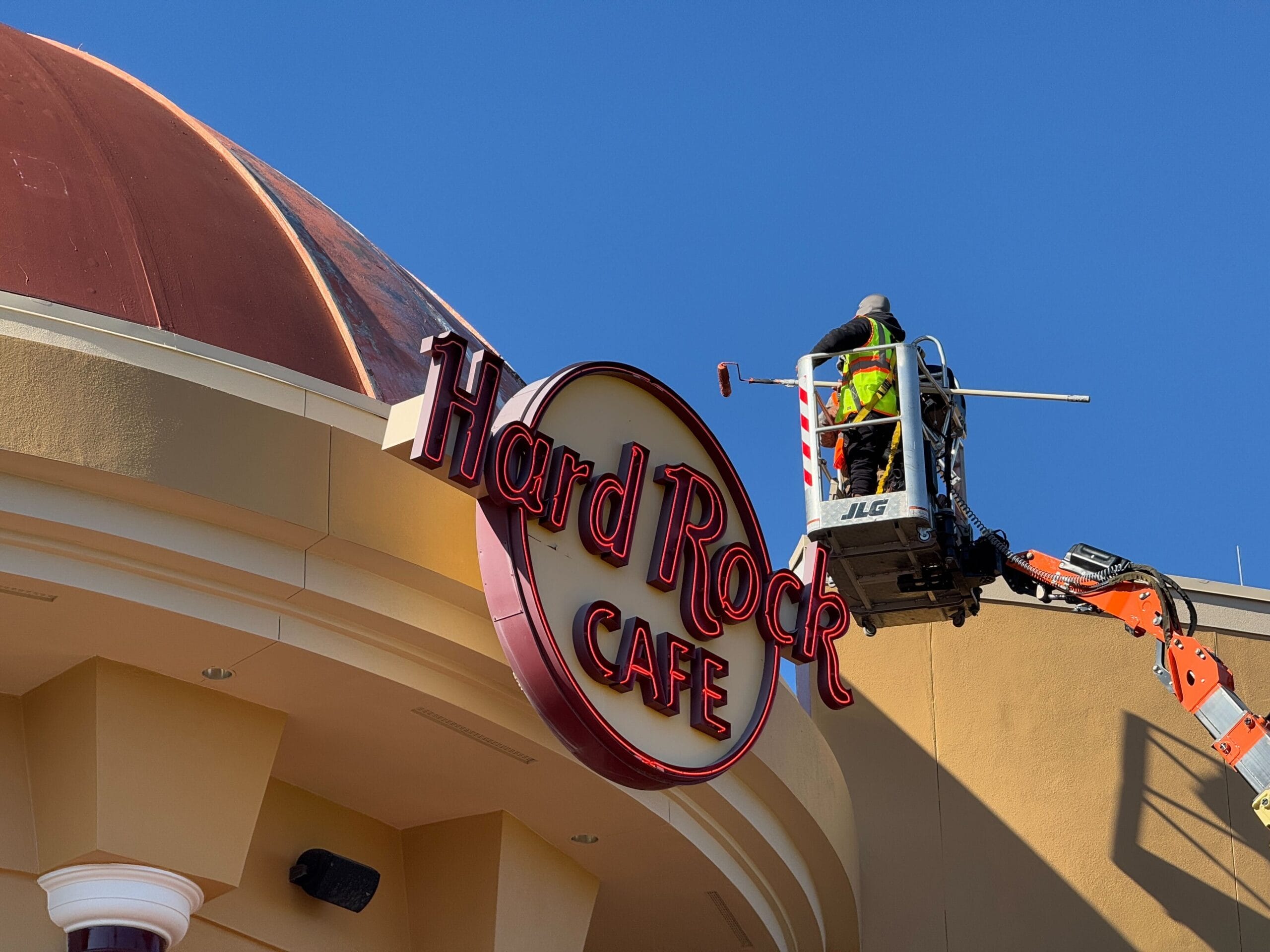 On a bright, clear day at Universal Orlando CityWalk, a worker on a lift carefully maintains the iconic Hard Rock Cafe sign atop the building, ensuring it stands out against the vivid blue sky.