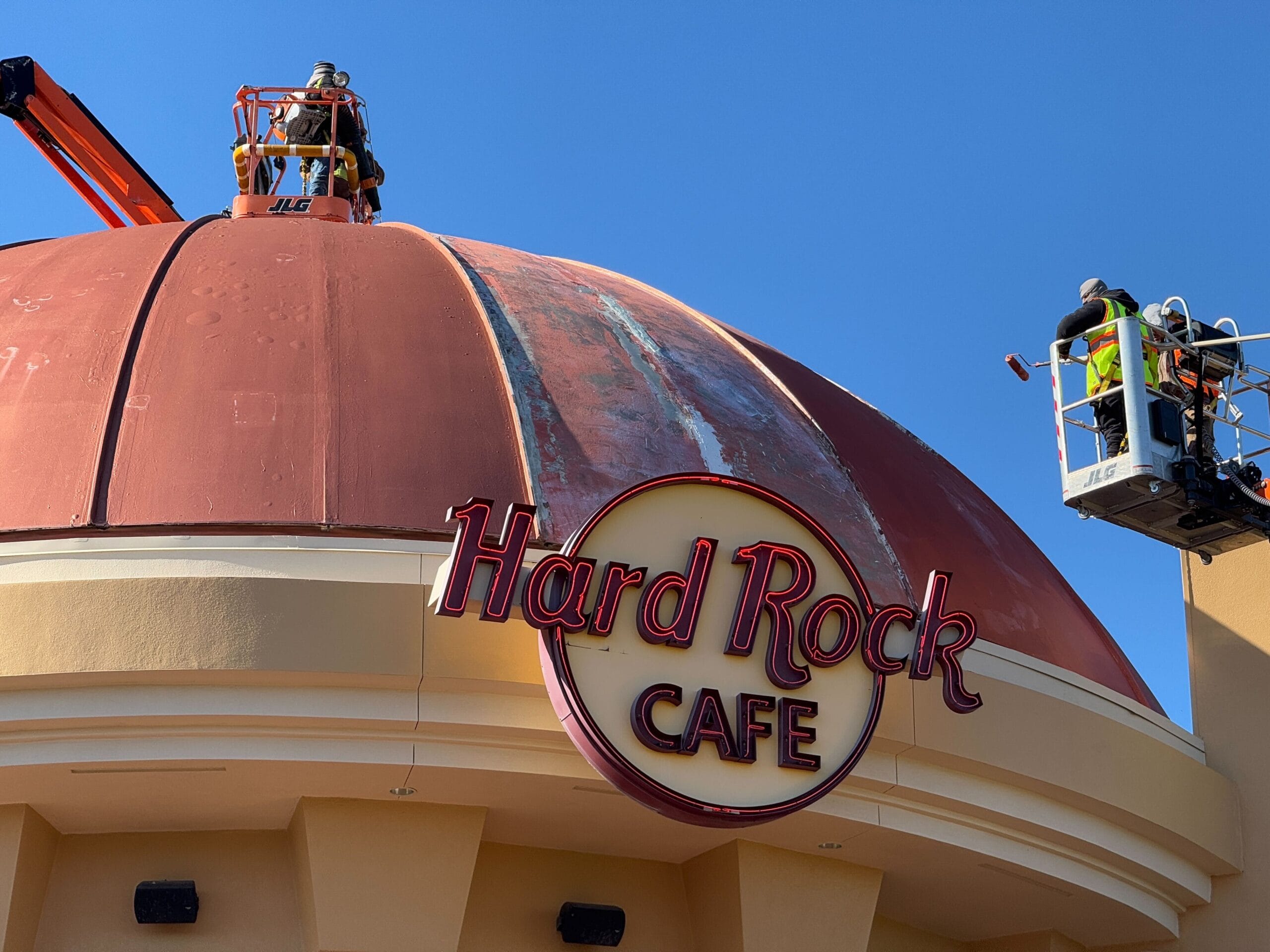 Workers on lift platforms conduct maintenance on the freshly repainted dome of the Hard Rock Cafe at Universal Orlando CityWalk, all under a clear blue sky.