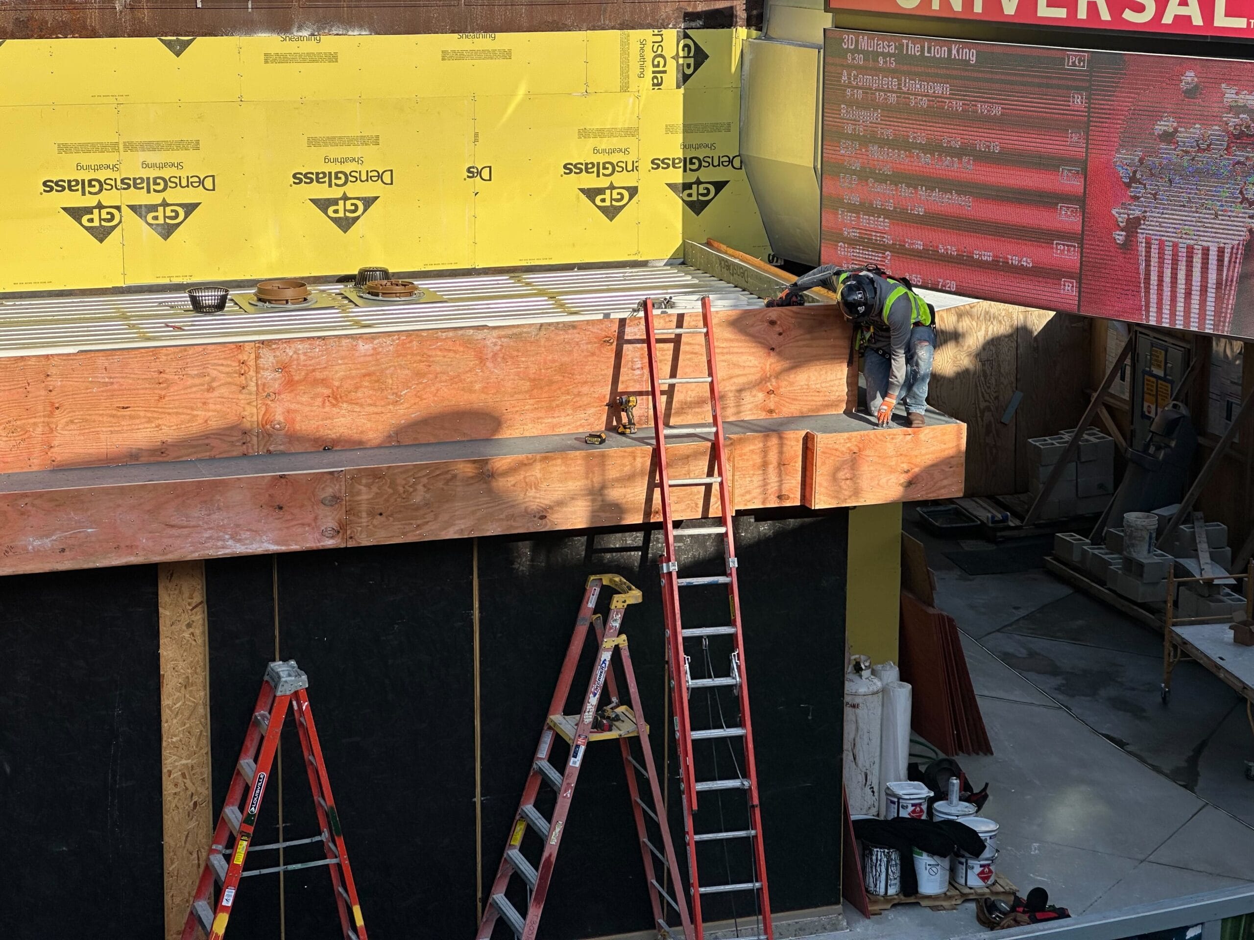Construction worker on a wooden platform beside a cinema, working near ladders. Movie posters and building materials are visible around the site.