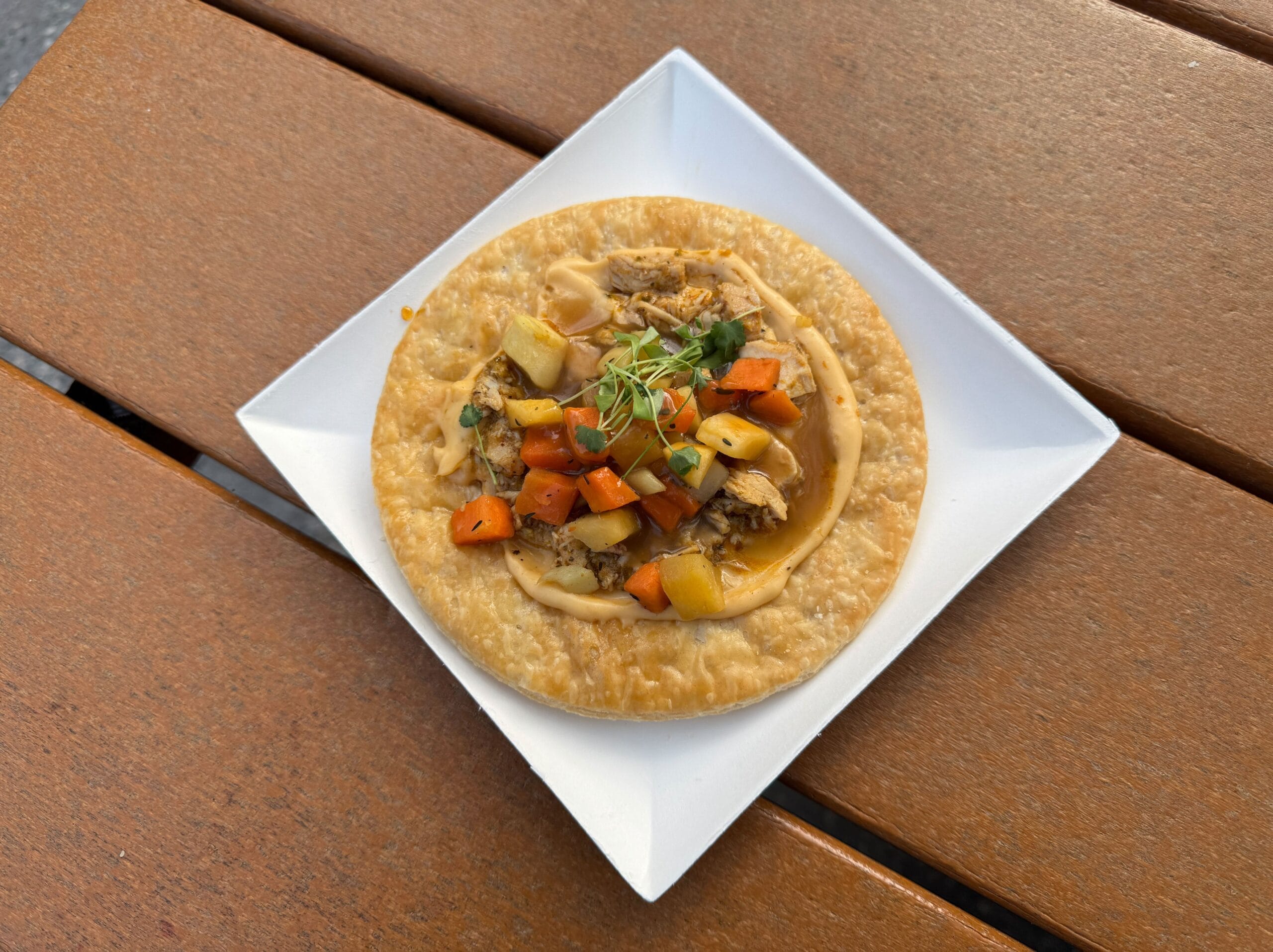 A savory pie topped with diced vegetables and microgreens rests on a square white plate, exuding a creative flair befitting the Festival of the Arts, elegantly displayed on a wooden table.